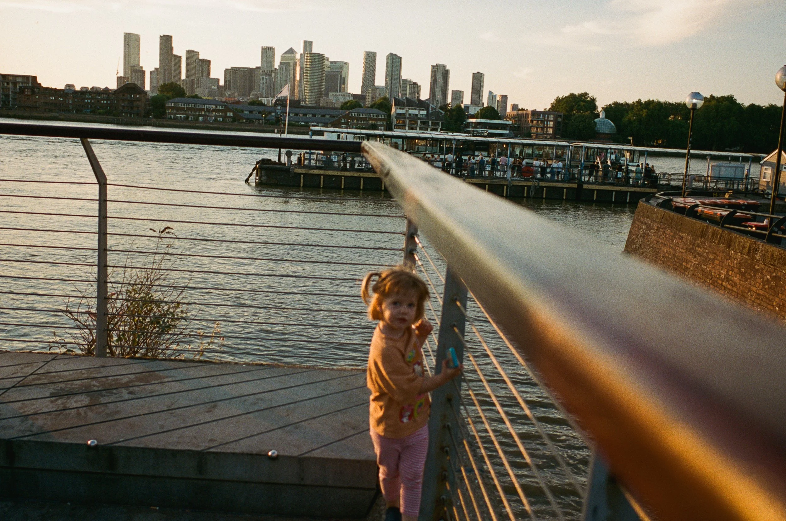 A young girl holding onto a railing on a waterfront walkway, with a city skyline in the background during sunset.