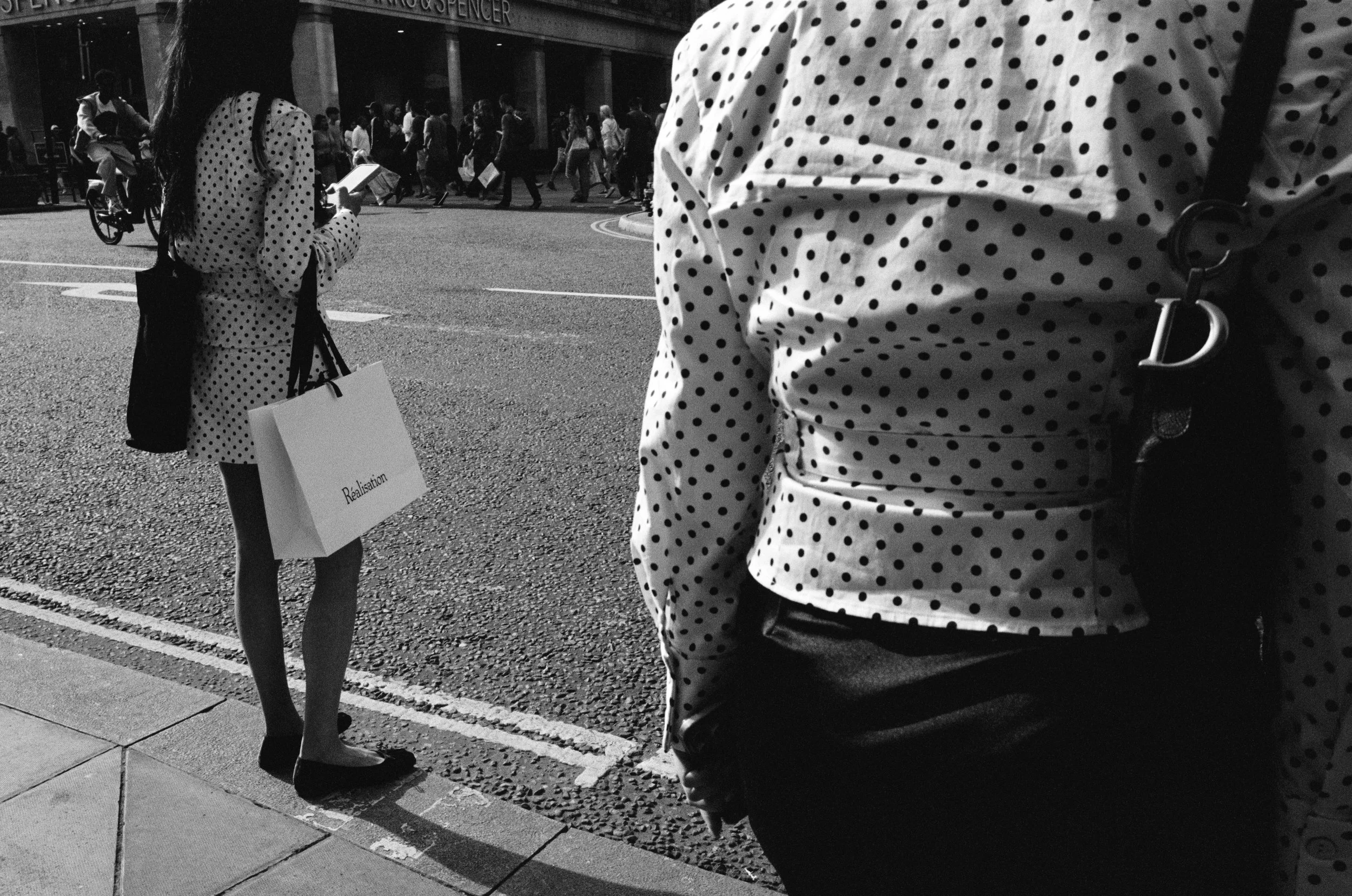 People walking across a busy city street, some carrying shopping bags, with a woman standing on the sidewalk looking at her phone, all dressed in polka dot clothing in black and white.