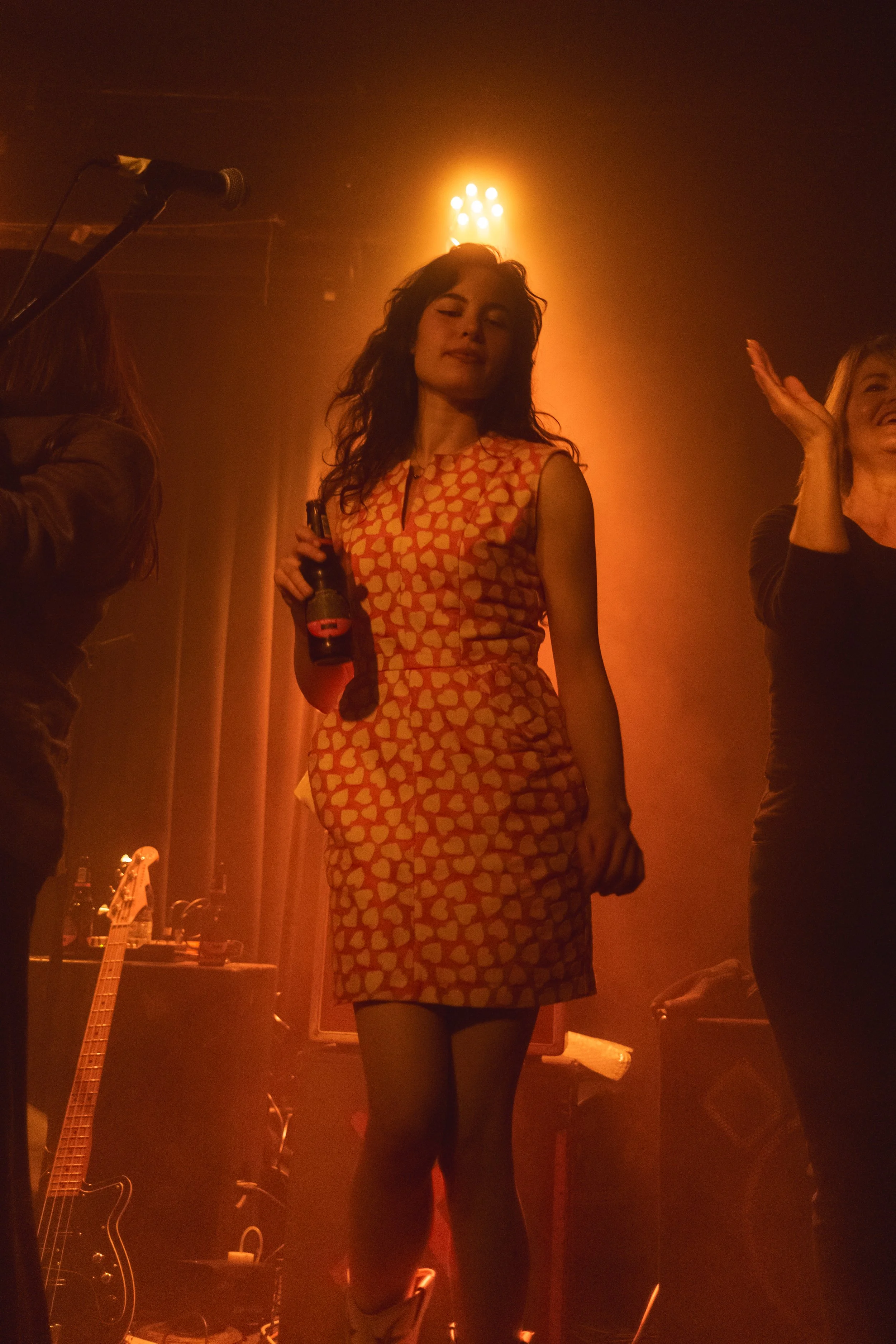 A woman with long, wavy dark hair, wearing a sleeveless dress with a red and yellow heart pattern, holding a bottle in her right hand, standing on stage with dim orange lighting and musical equipment in the background.