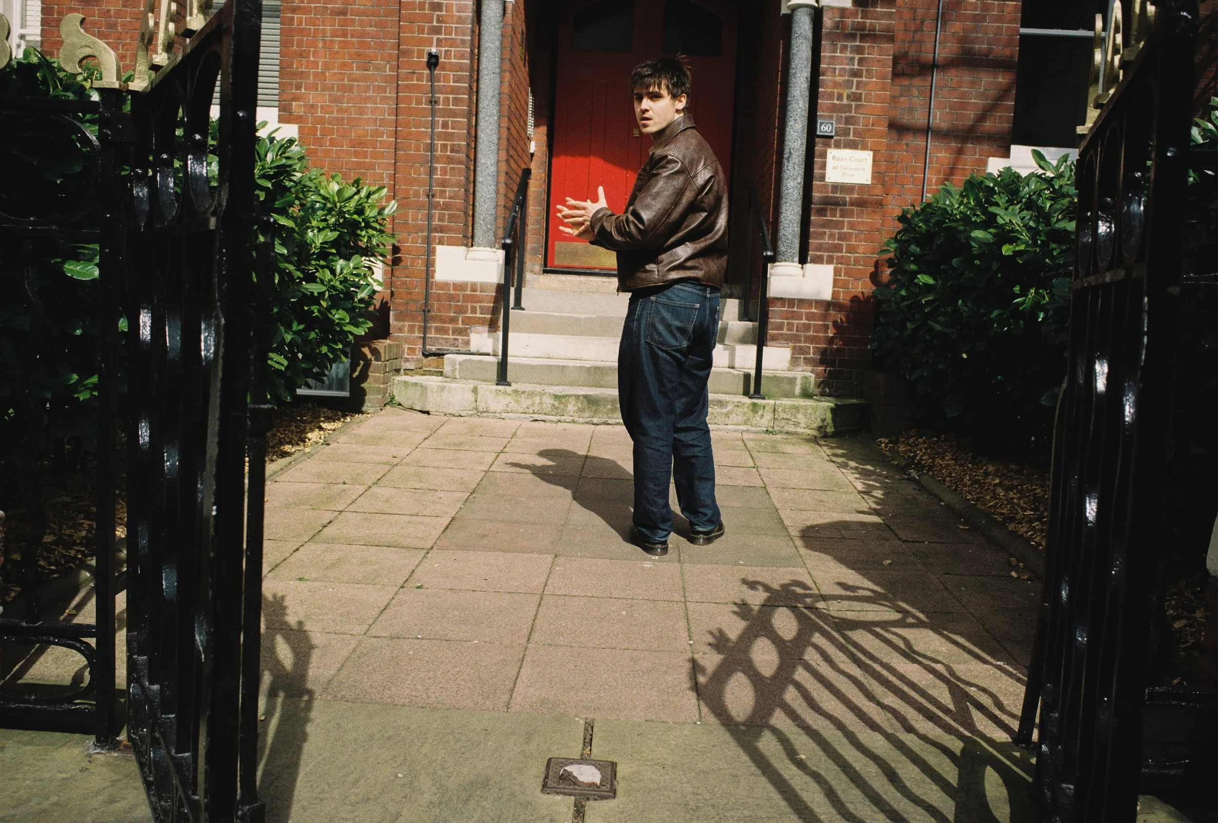 A young man in a brown leather jacket and jeans is standing outside on a sidewalk near a brick building with a red door. He is clapping his hands and looking to the side. The scene is framed by black iron gates and green bushes.