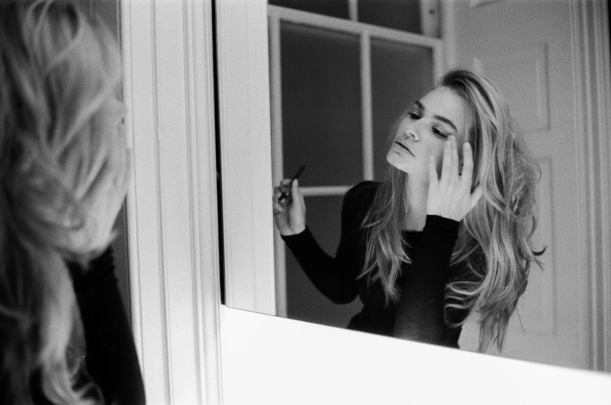 A woman with long, wavy hair is looking into a mirror, touching her face with one hand and holding a makeup brush in the other. She appears to be applying makeup in a bedroom or dressing area.