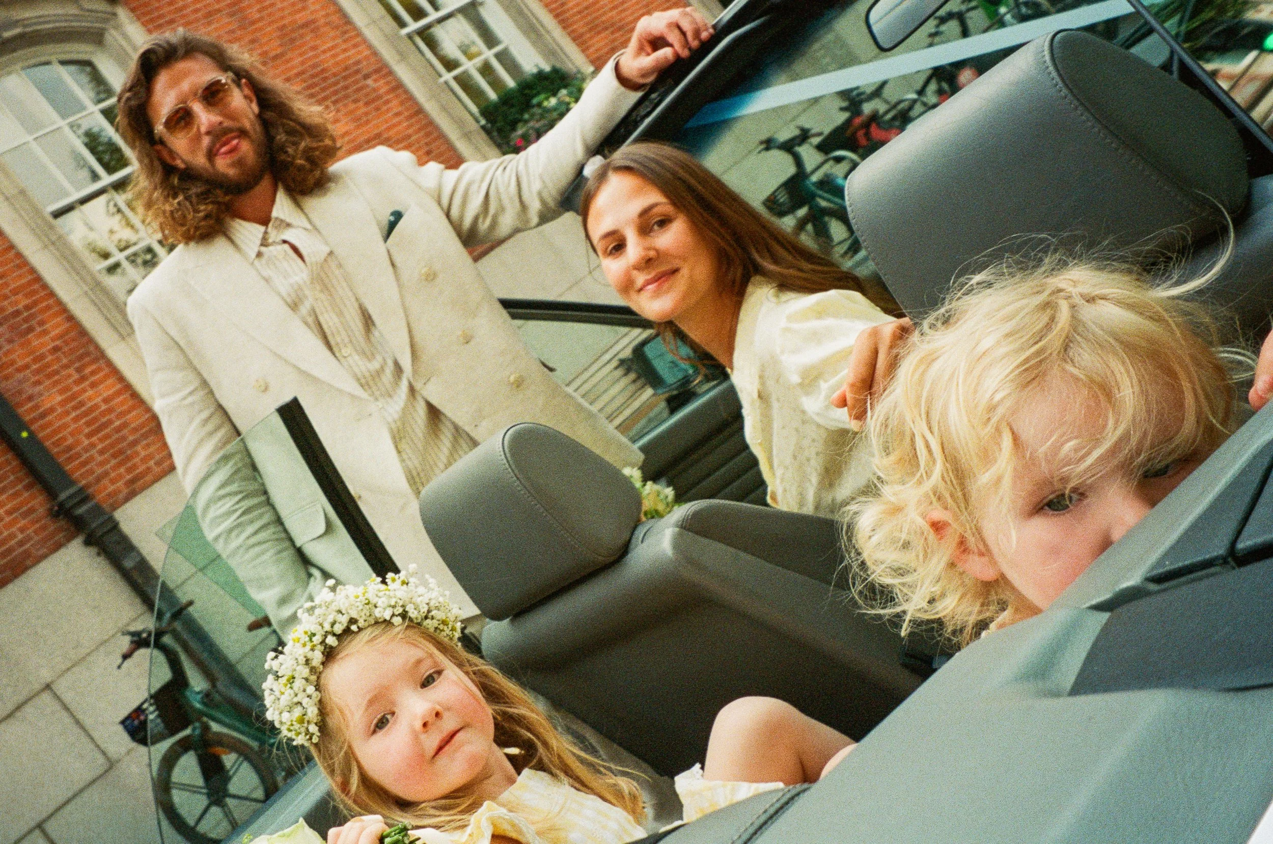A man with long hair and sunglasses, dressed in a white suit, standing next to a woman and a child in a convertible car. The woman has shoulder-length hair and is smiling, while the child has blonde hair and wears a flower crown, looking at the camer