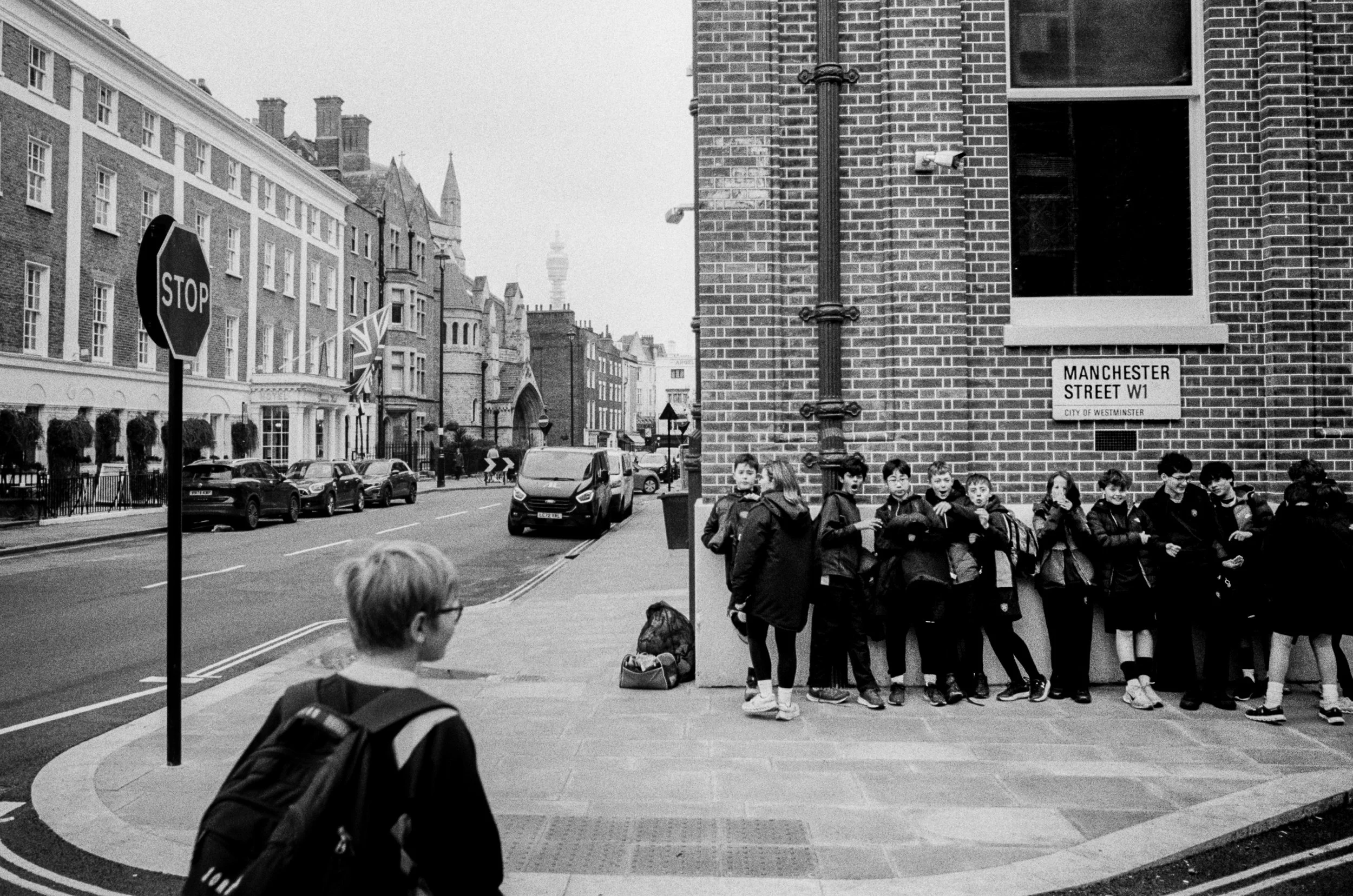 A group of school children standing in line on the sidewalk on Manchester Street in Westminster, London, with a row of traditional brick buildings and the TV Tower in the background.