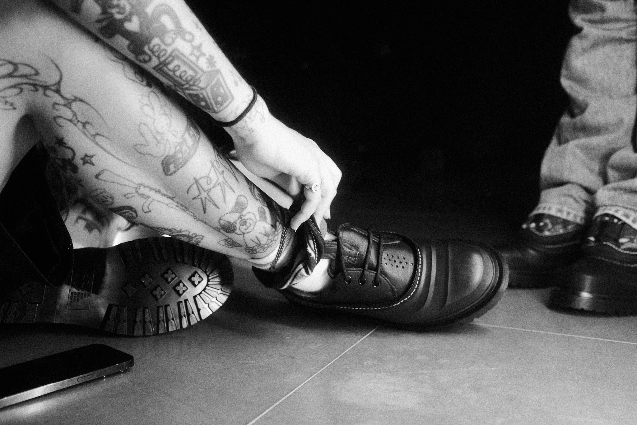 Close-up black and white photograph of a person's tattooed arm wearing a black T-shirt, a black bracelet, and a ring, resting on a flat surface near a pair of black boots with rounded toes and laces. The background shows a patterned rug and part of a