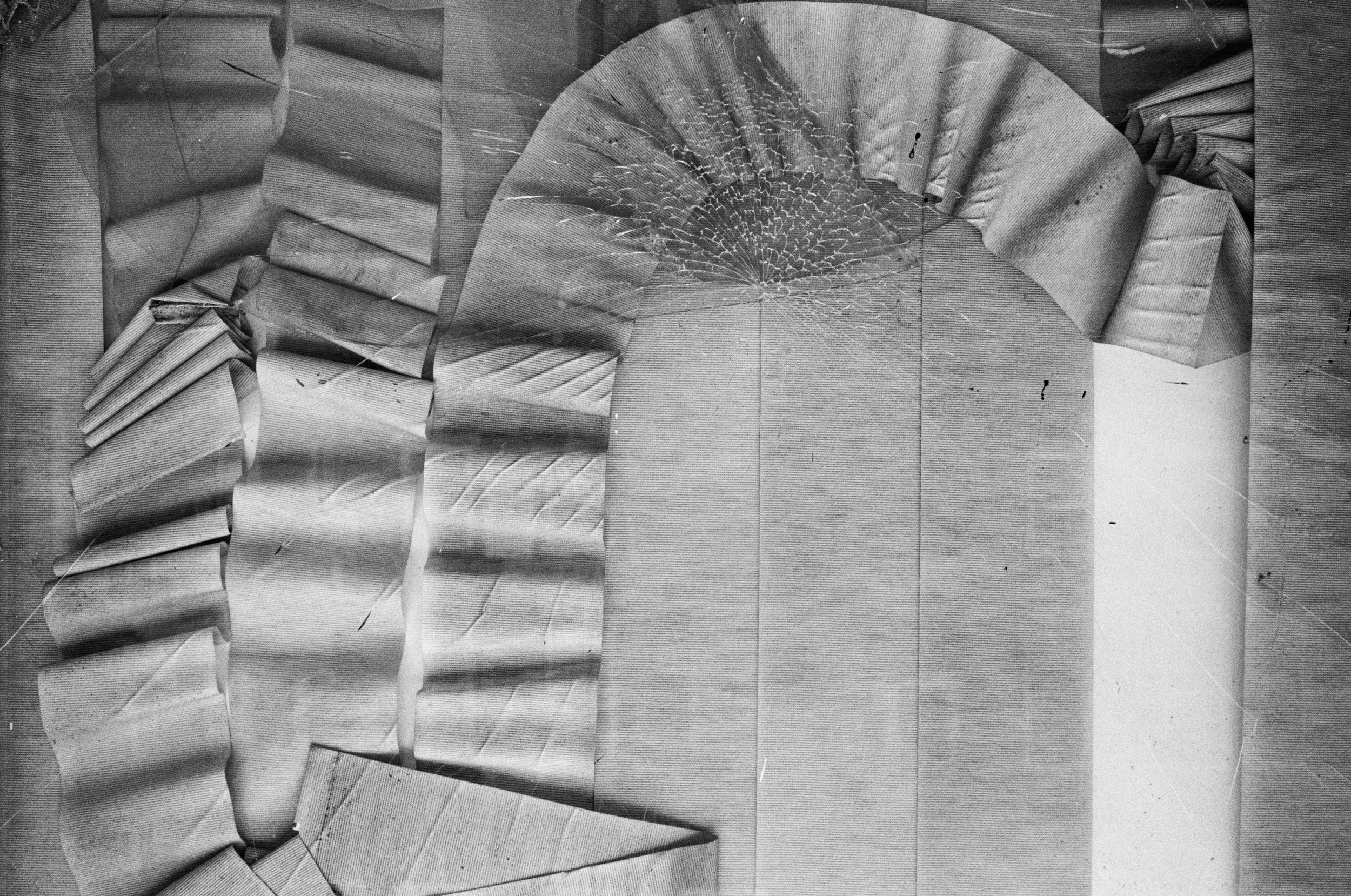 Black and white image of a large, broken ceiling fan with multiple blades, some bent or broken, against a background of textured ceiling tiles.