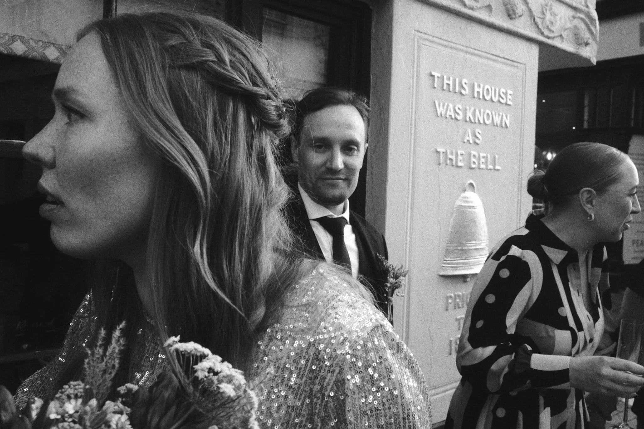 Black and white photo of three women and a man in formal attire, sitting at a table in a restaurant or event space. One woman in the foreground has long wavy hair and is wearing a sequined top. The man in the middle is smiling and wearing a suit and 