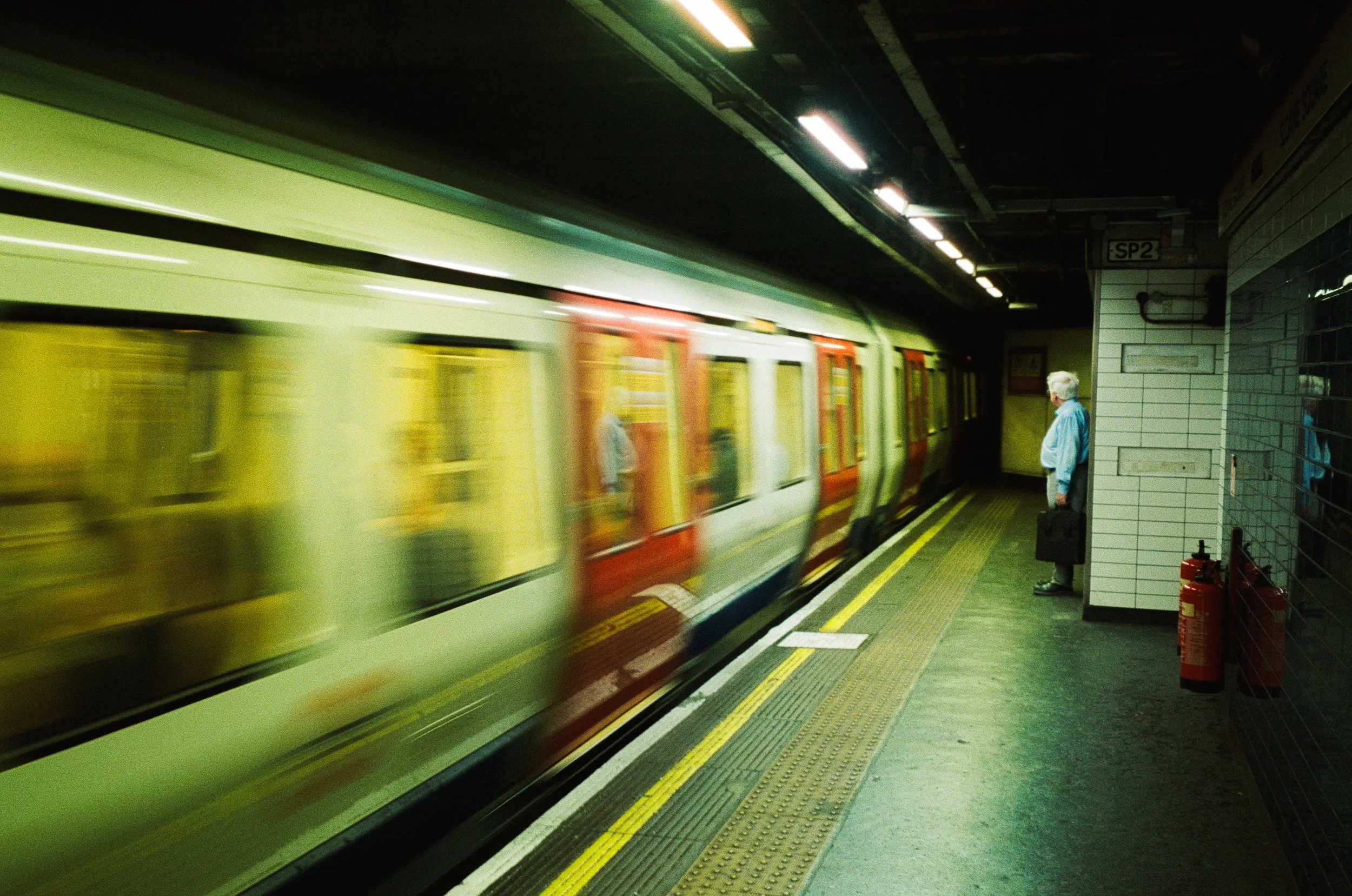 A subway station platform with a train arriving and a man standing on the platform. The train is yellow with red and white accents, and the setting is underground with tiled walls and fire extinguishers nearby.