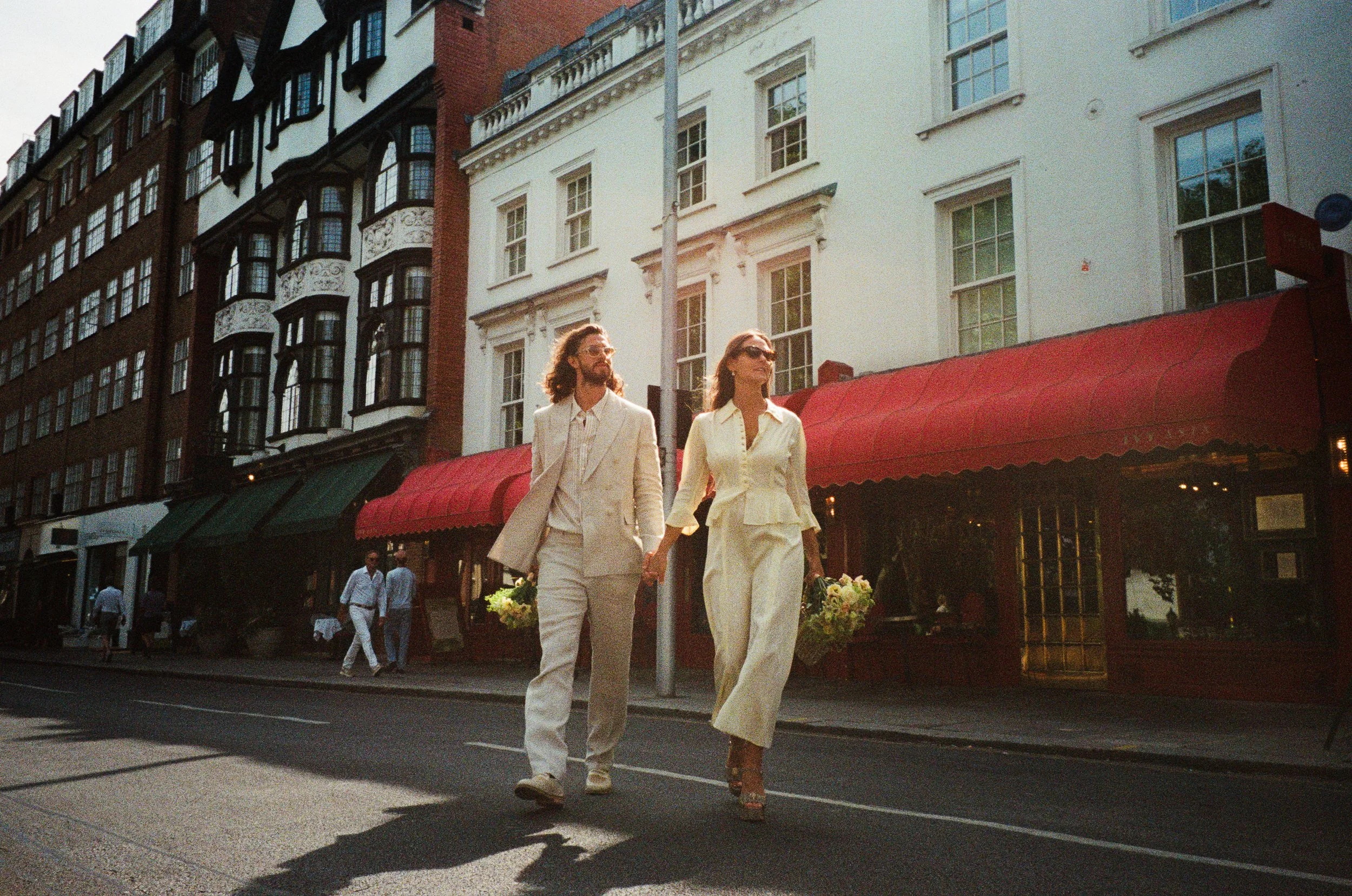 A couple dressed in white walking hand in hand on a city street during sunset, carrying bouquets of flowers.