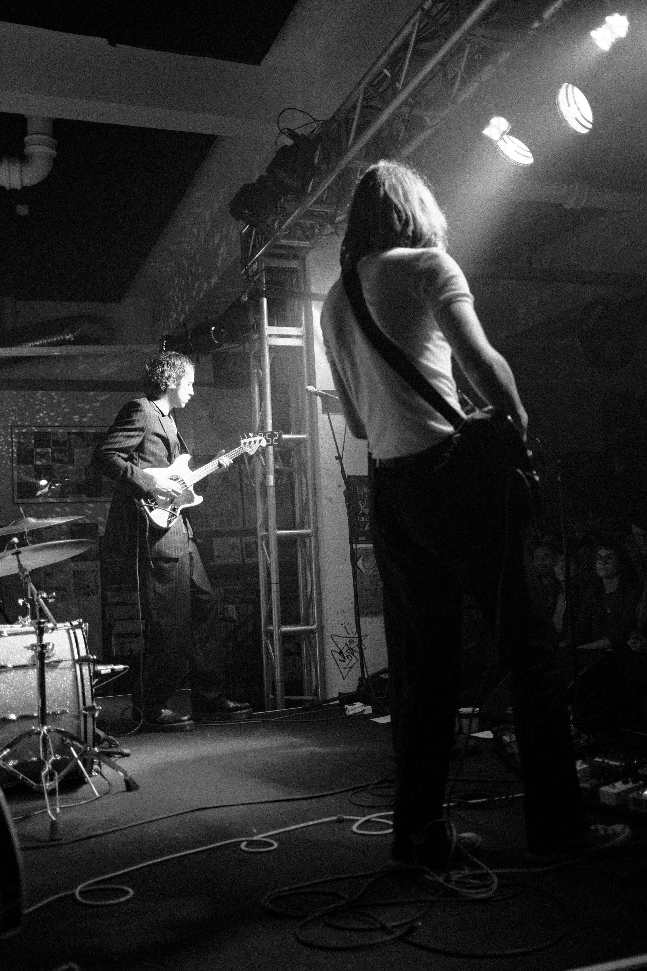 Black and white photo of a live music performance with two male musicians on stage. One is playing an electric guitar, and the other appears to be singing or speaking into a microphone. There are stage lights and equipment along with cables on the fl