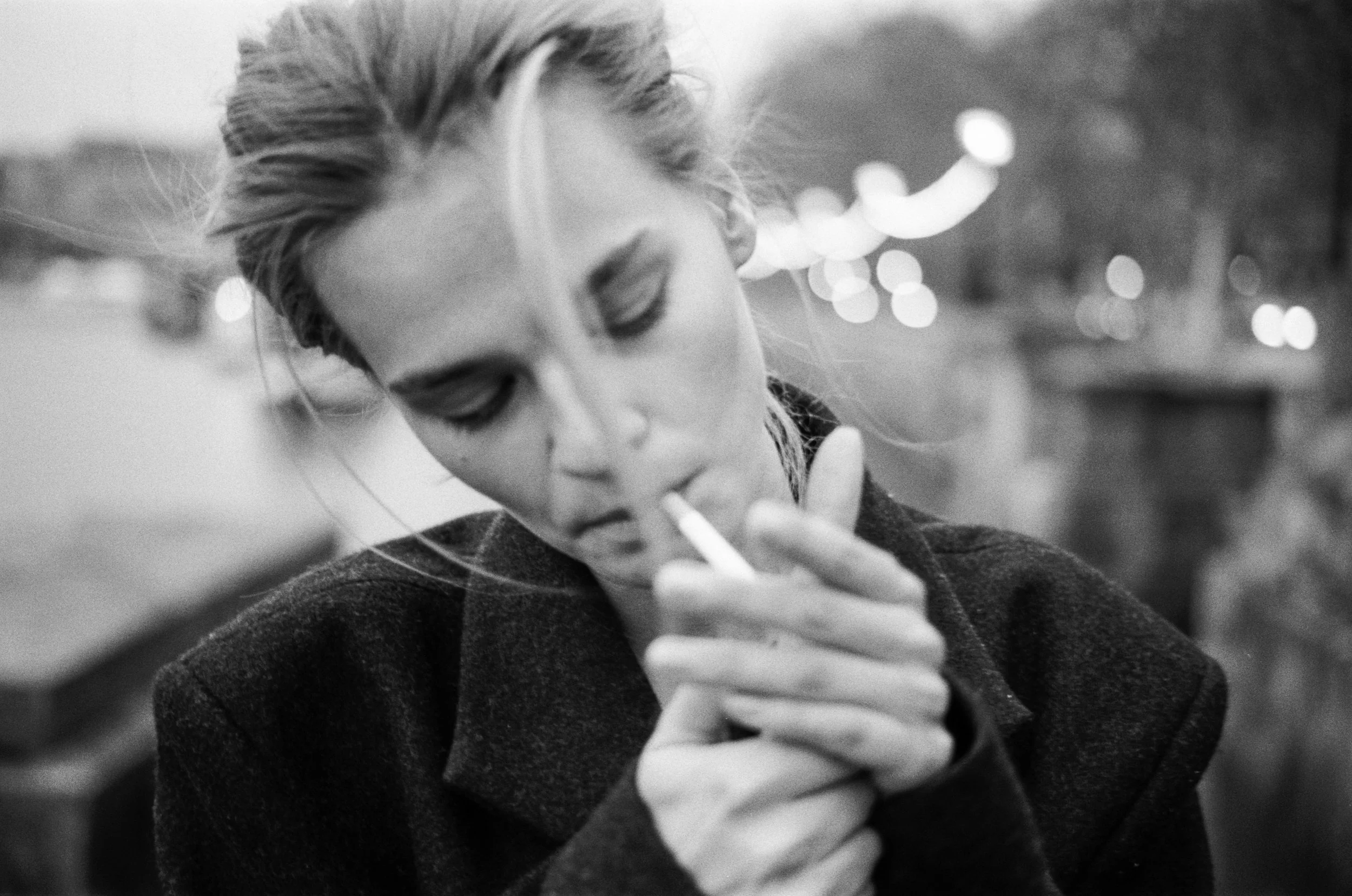 A woman with blonde hair styled in a bun, wearing a dark coat, looking down and smoking a cigarette outdoors at night with blurred streetlights in the background.