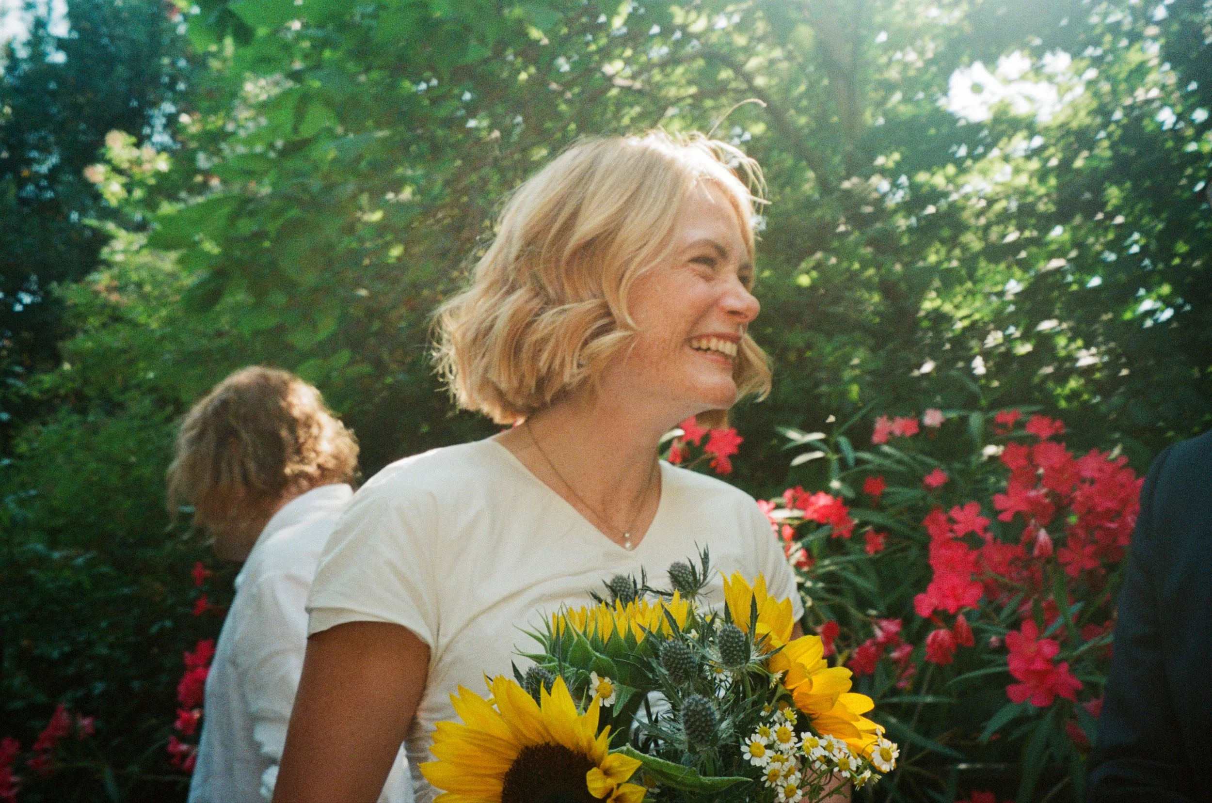 A woman with blonde hair smiling and holding a bouquet of sunflowers, standing outdoors amidst colorful flowers and green foliage.