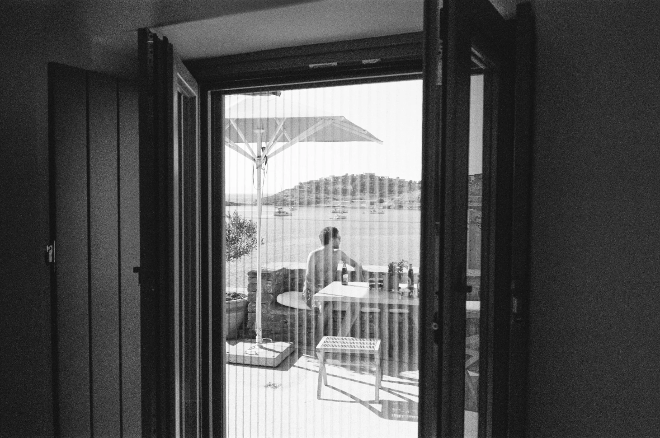 A person standing outside on a balcony or patio area, with a large umbrella and a table, overlooking a body of water with boats and hills in the background, seen through an open sliding door.