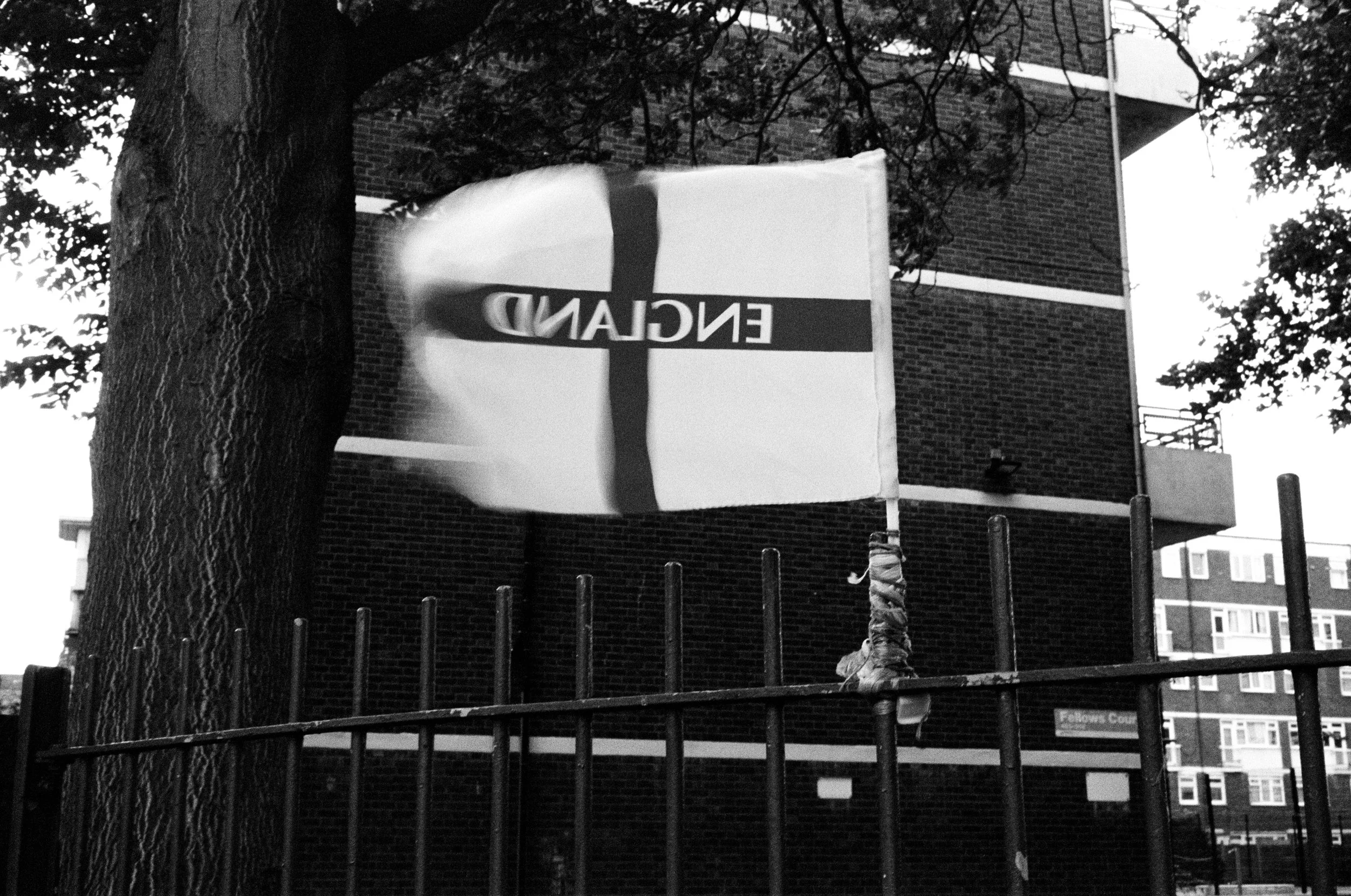 Black and white photo of a street scene with a large tree in the foreground, a building with brick walls, a flagpole, and a sign that is partly cut off. The sign has the word "ENGLAND" visible in reversed letters.