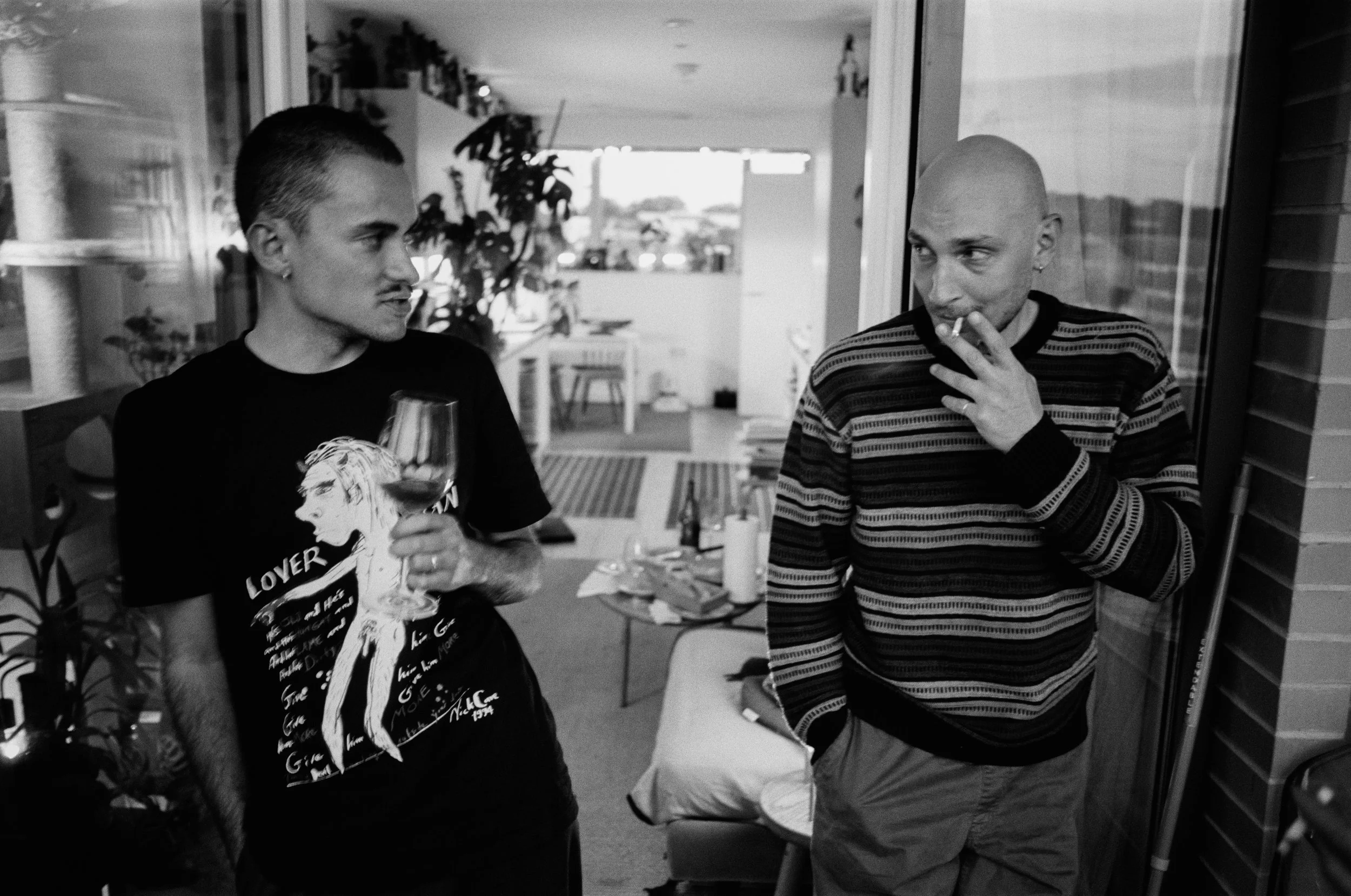 Two men engaged in conversation indoors; one holding a glass of wine, the other smoking a cigarette. Both men have shaved heads and are dressed casually.
