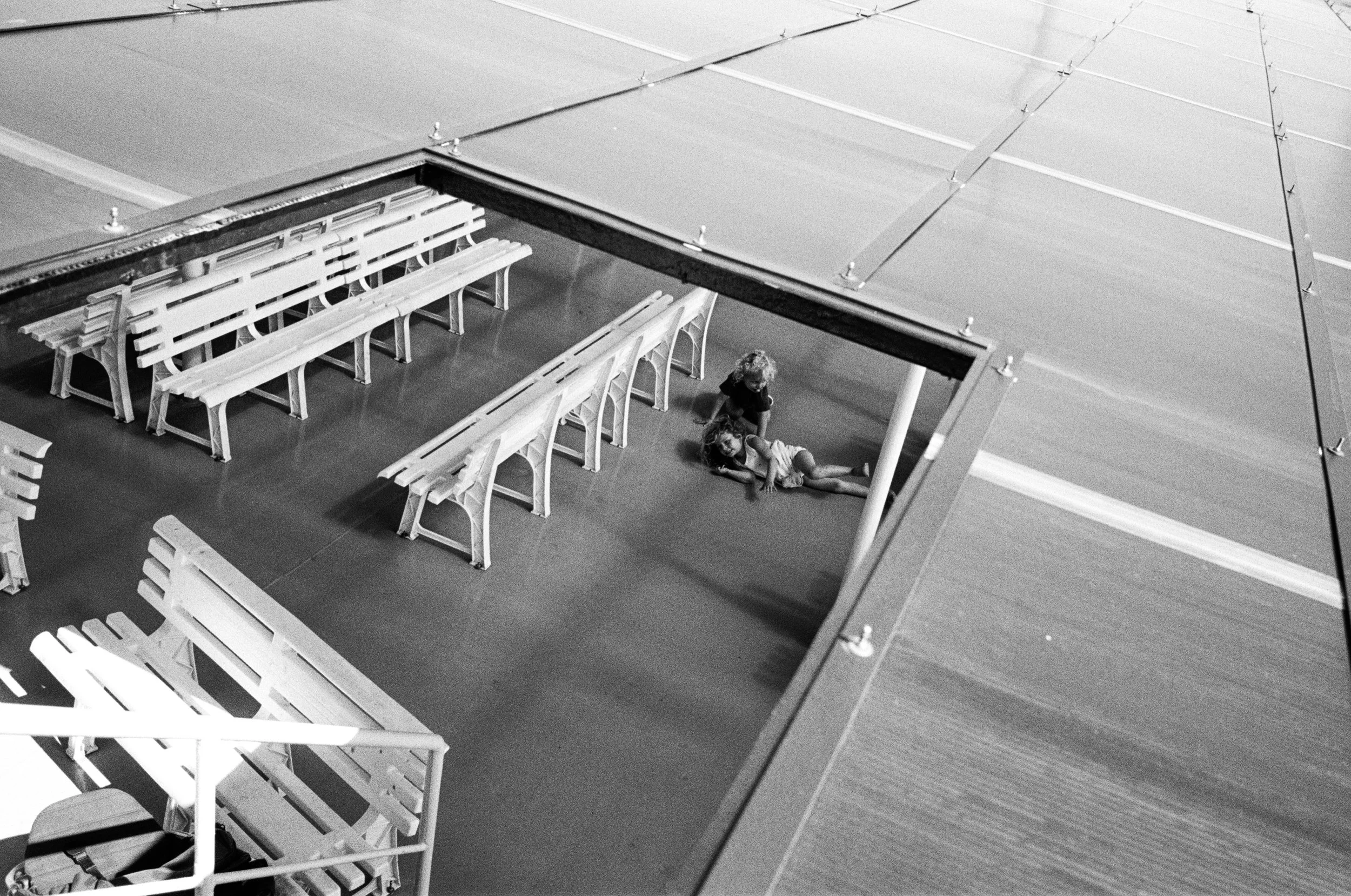 Two children sitting on the floor under a roof, with benches and tables around them, in a covered outdoor area.