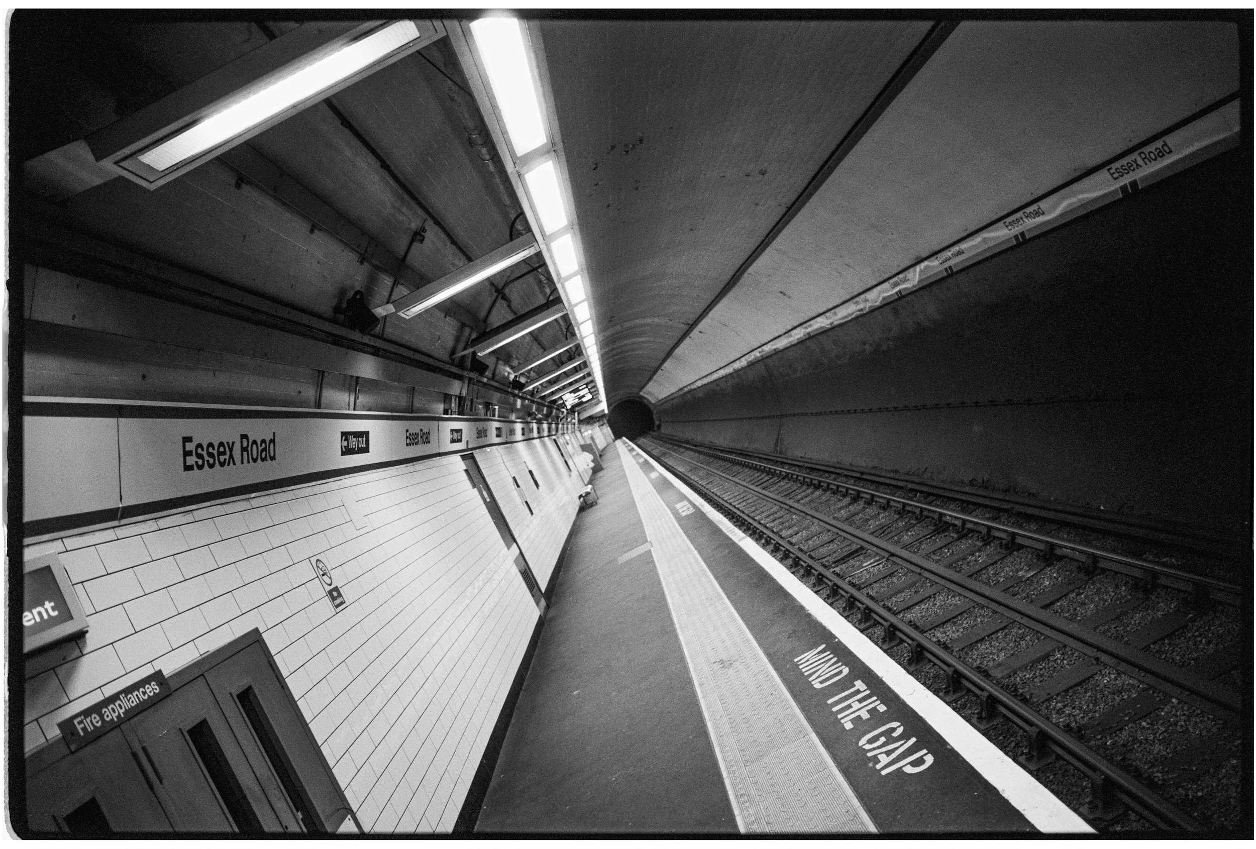 Empty underground subway station platform, labeled 'Essex Road,' with bright lighting and tracks visible on the right side.