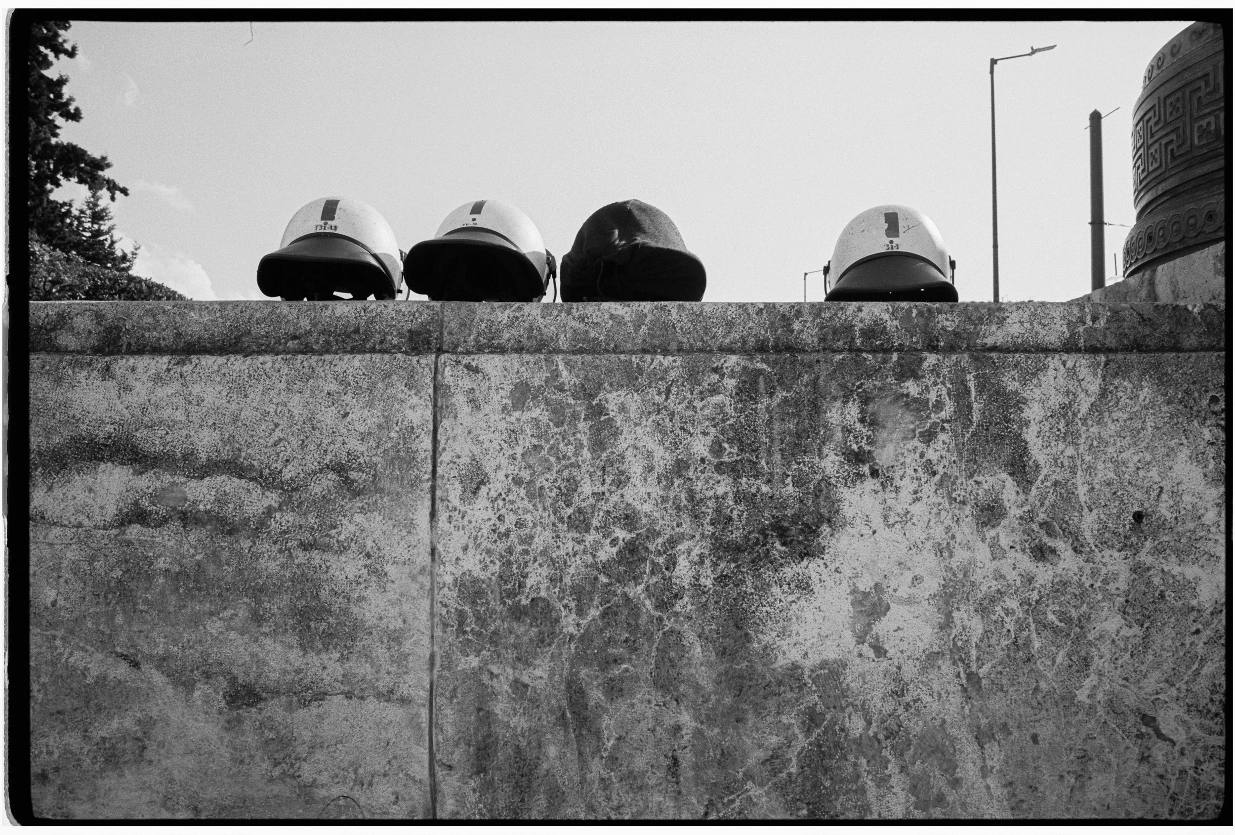 Four motorcycle helmets on a stone railing, with a person's head in the middle, outdoors under a cloudy sky, with trees and streetlights in the background.