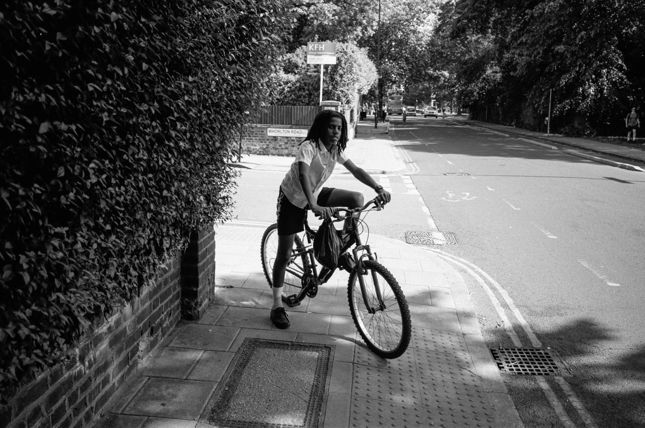 A person with dreadlocks riding a bicycle on a city sidewalk next to a brick wall and a hedge. The street in the background has trees, cars, and a bus. A sign reads "KFH" and another reads "Whorton Road." The scene is in black and white.