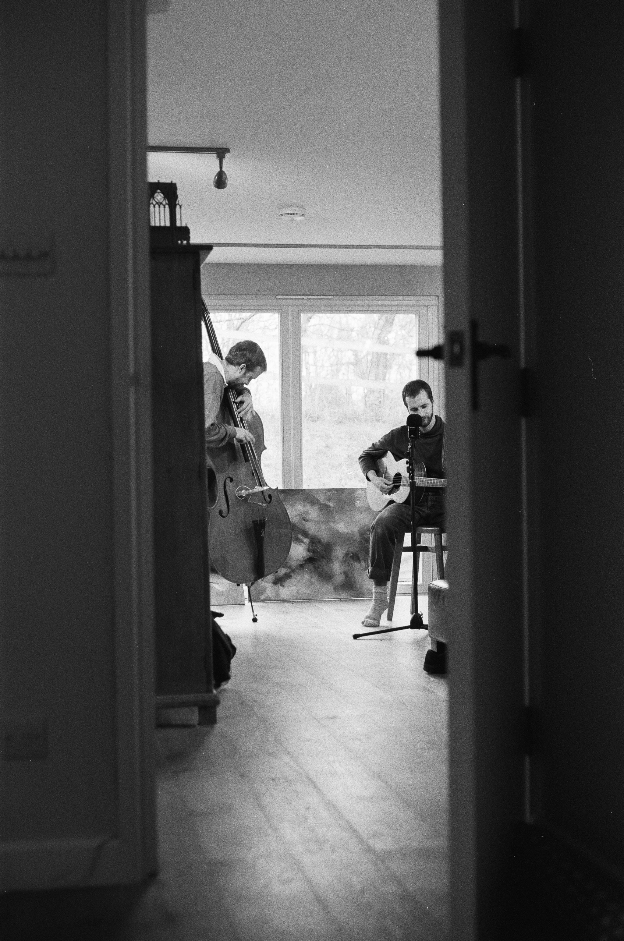 Two men playing musical instruments, a double bass and an acoustic guitar, in a room with large windows. One man is standing and the other is sitting on a stool, both are singing into a microphone.