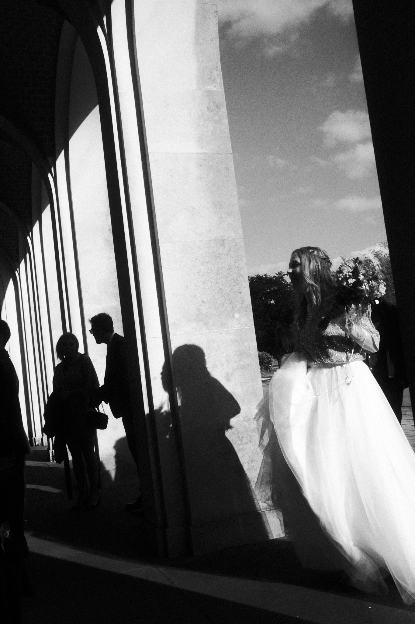 A woman in a wedding dress holding a bouquet of flowers, standing outside near a modern building with large windows and columns. Shadows of people are cast on the ground.