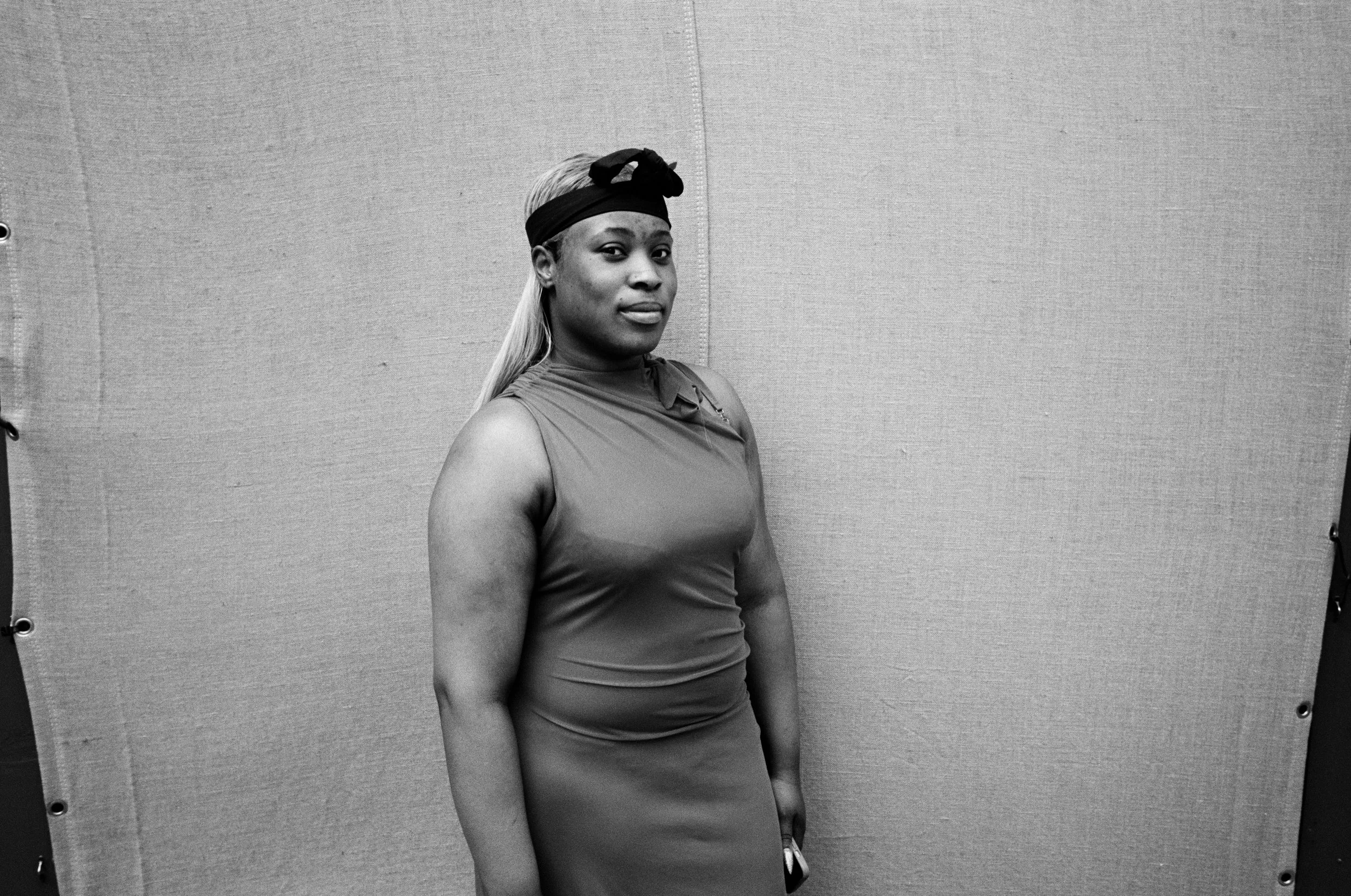A black and white photo of a woman with long, platinum blonde hair wearing a black headband and a sleeveless top. She is standing in front of a plain, textured wall.