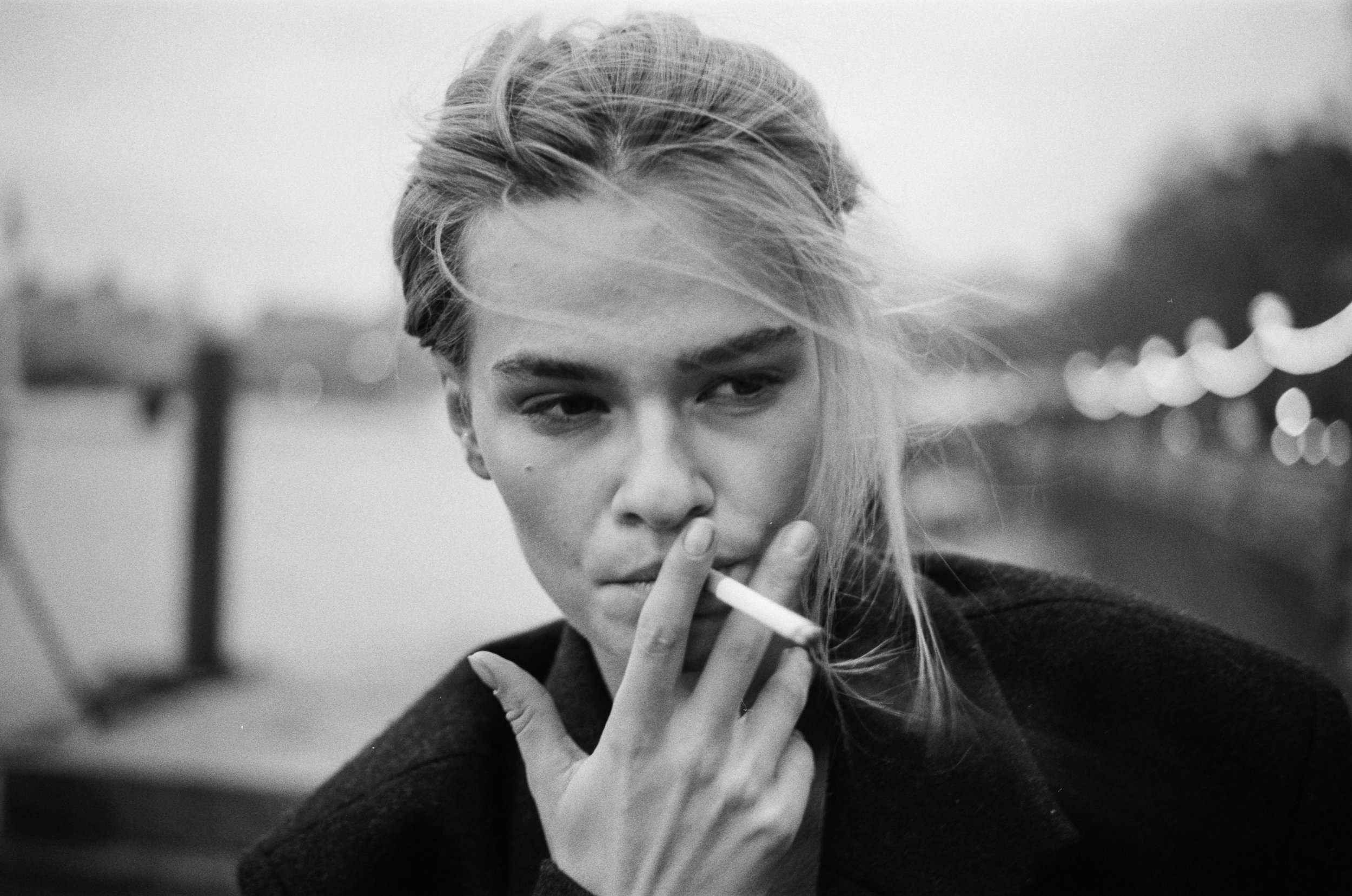 A black-and-white close-up of a young woman with light hair, holding a cigarette in her hand with her lips slightly parted and her eyes looking downward.