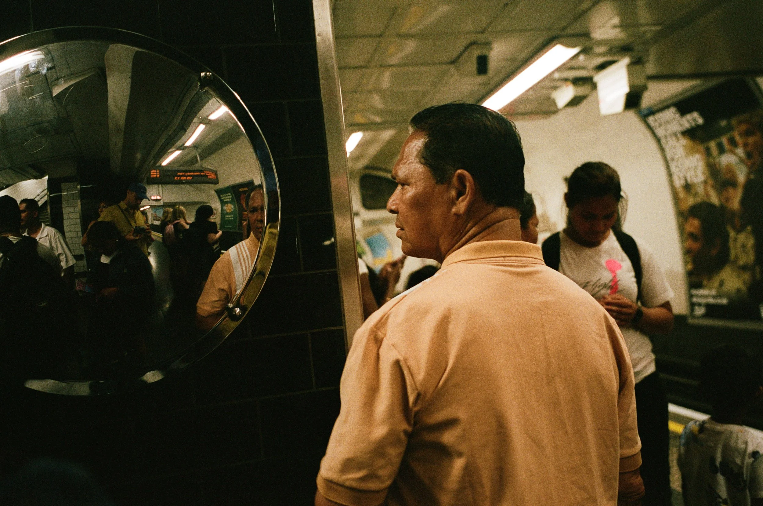 A man with dark hair wearing a peach-colored shirt stands facing away from the camera in a subway station, reflected in a curved mirror on the wall. Other passengers are visible in the background, some using their phones, with an underground subway p