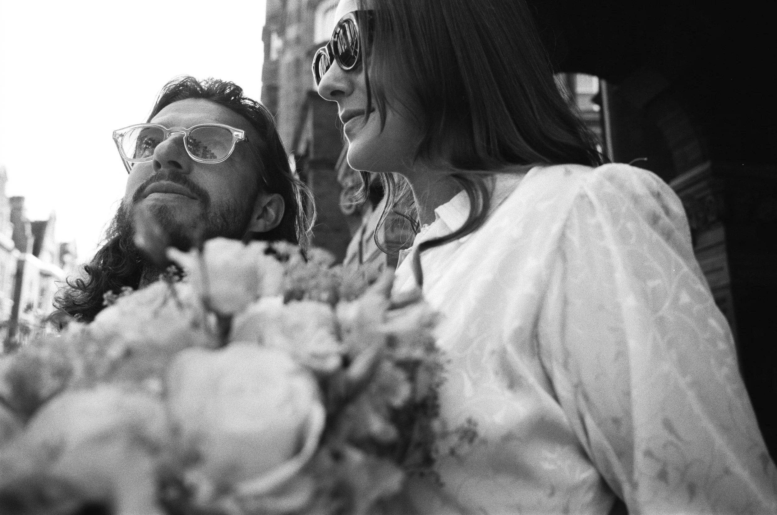 A black and white photo of a man and woman sitting outdoors, wearing sunglasses, with food on the table in front of them.