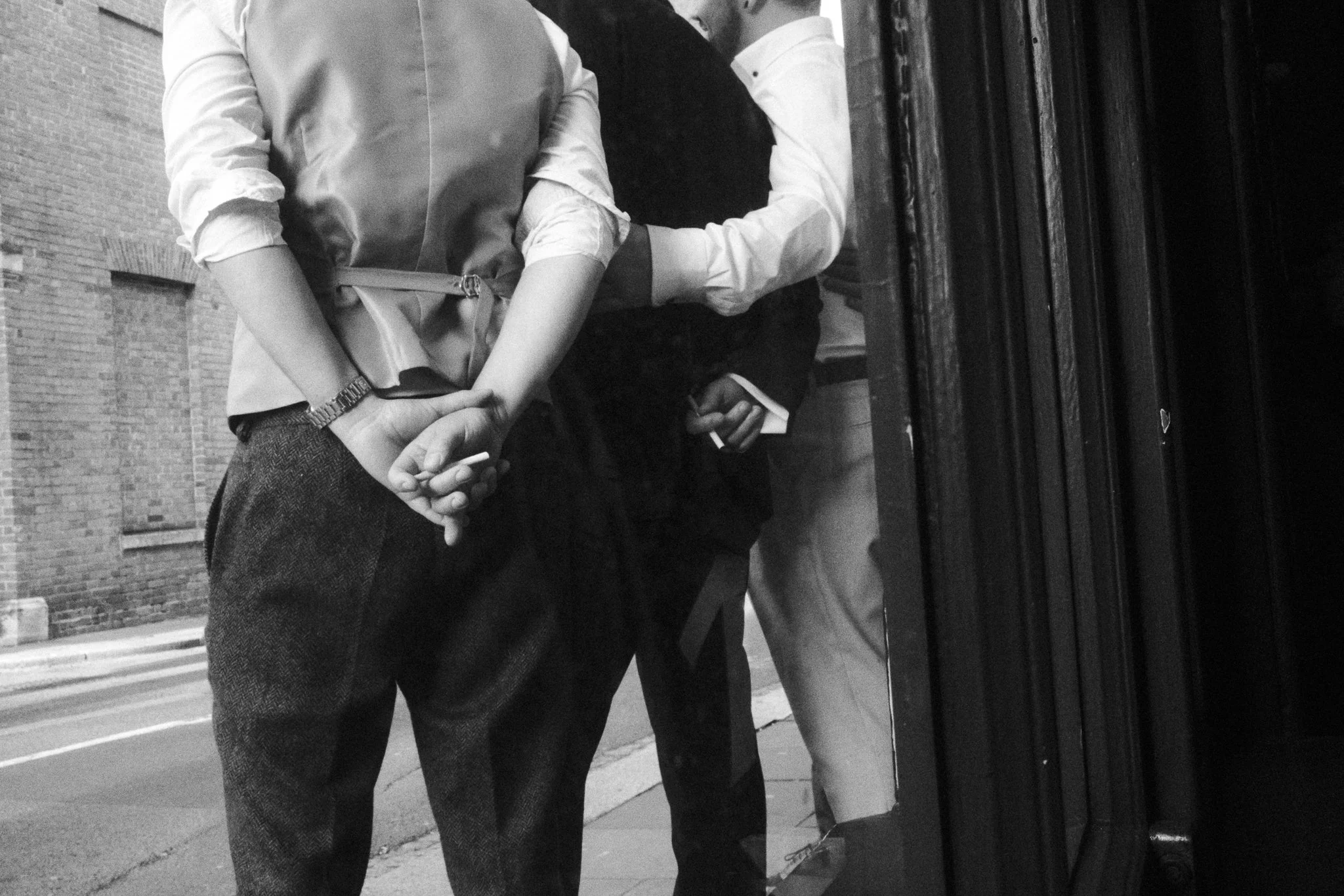 Two people holding hands on a city sidewalk, seen in black and white, with an industrial brick building in the background.