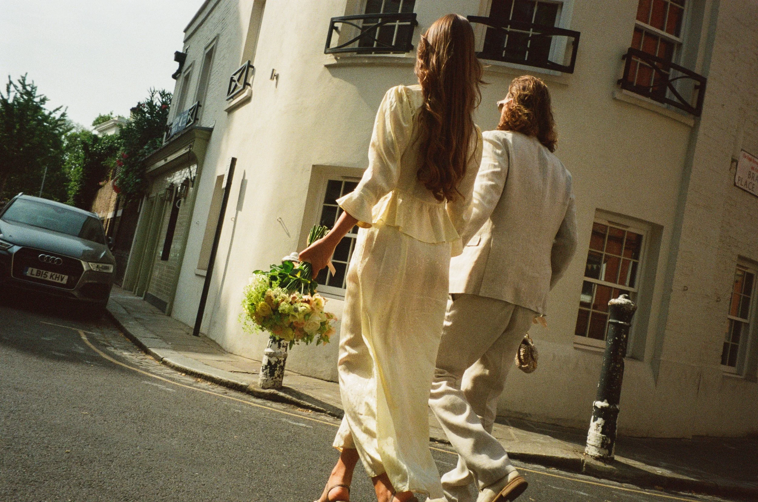 Two women dressed in light-colored clothing walking down a street, one carrying a bouquet of flowers, with residential buildings and cars in the background.