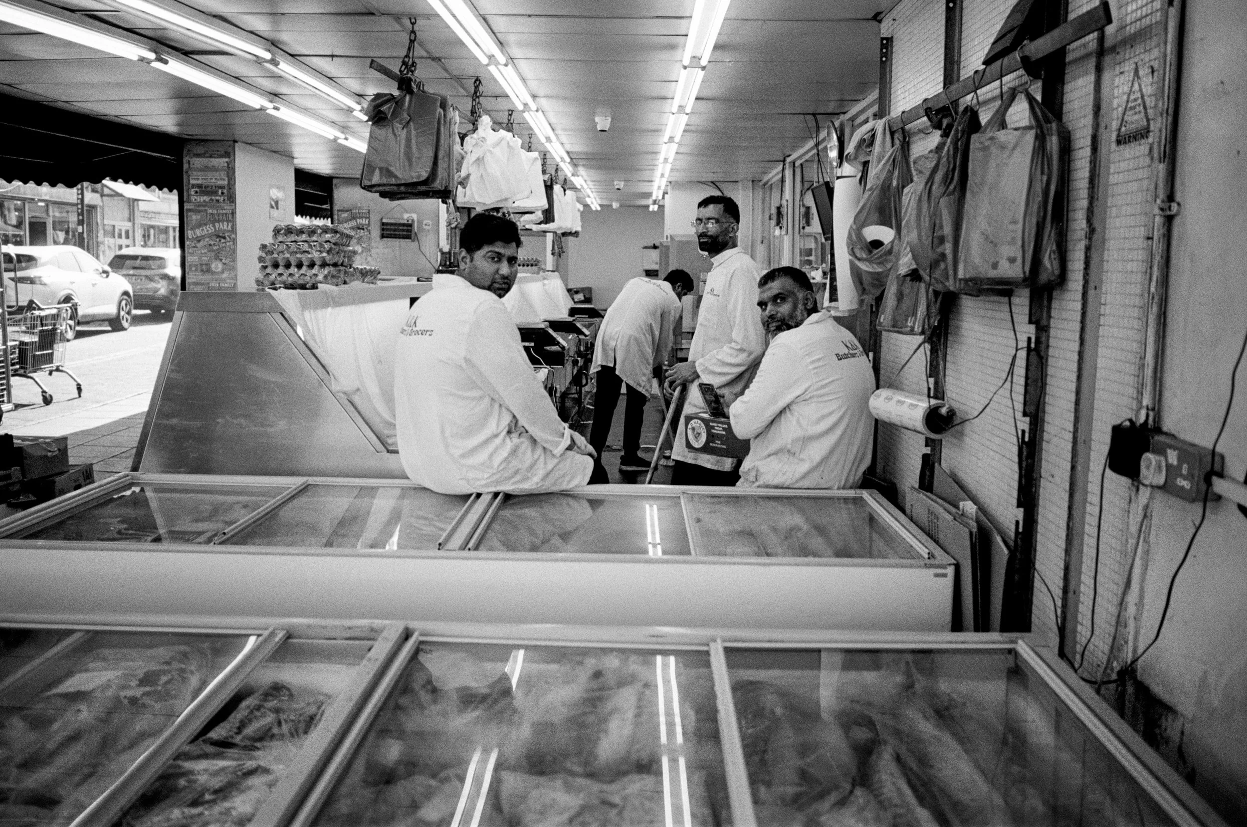 Four men in white coats sitting at a counter inside a grocery store, with shelves and bags hanging in the background.