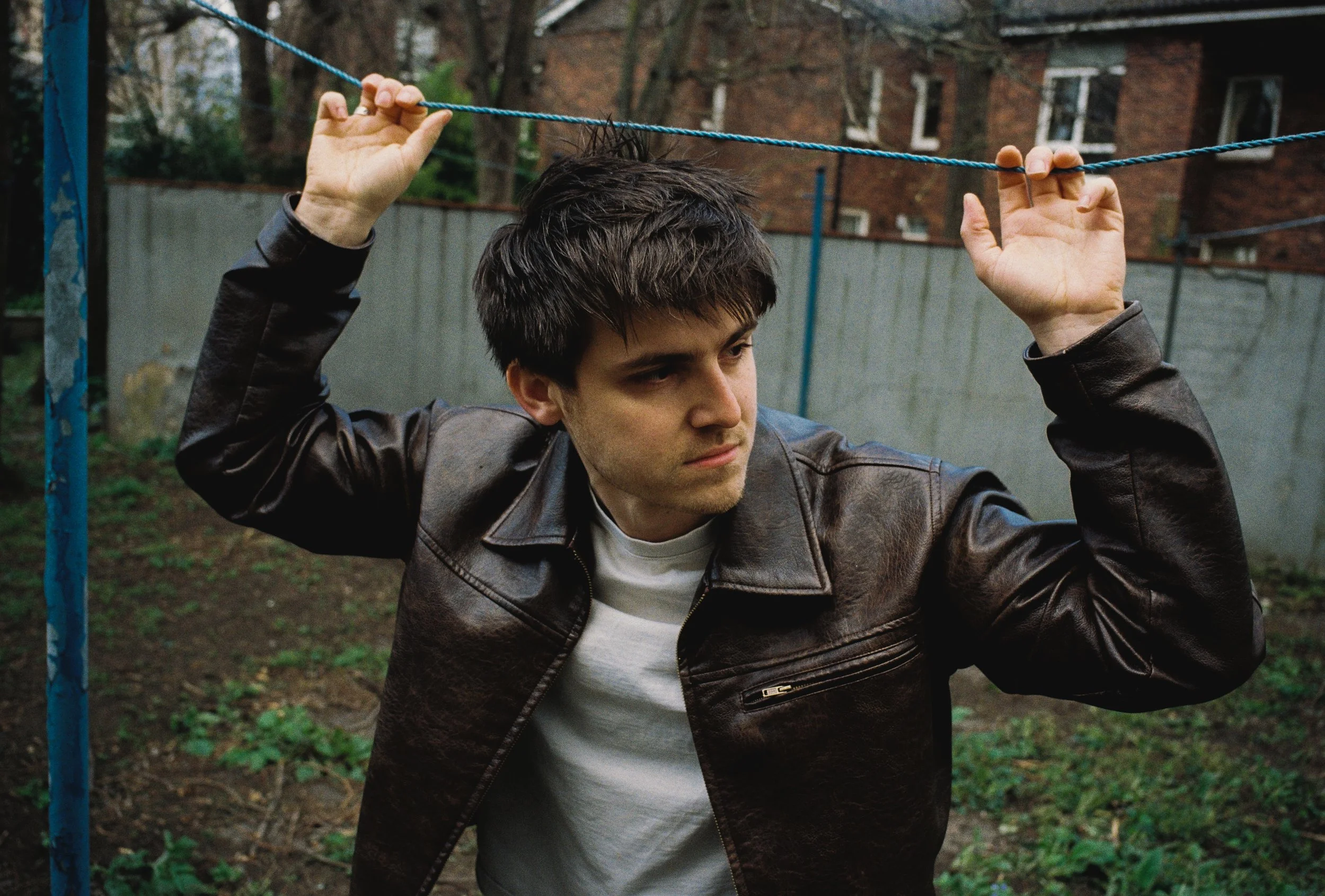 Young man in a leather jacket hanging from a blue rope in an outdoor setting.