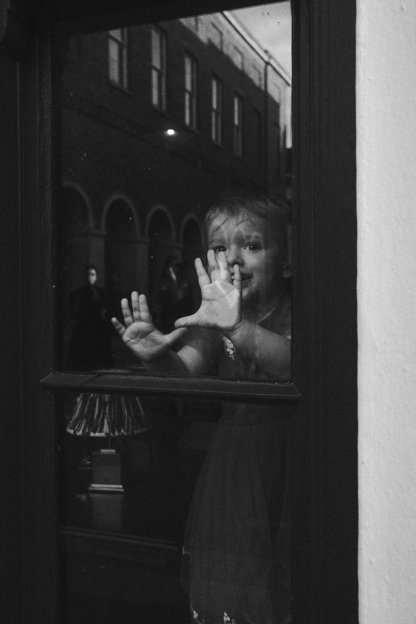 A young girl is standing behind a glass window, pressing her hands and face against the glass and smiling at the camera.
