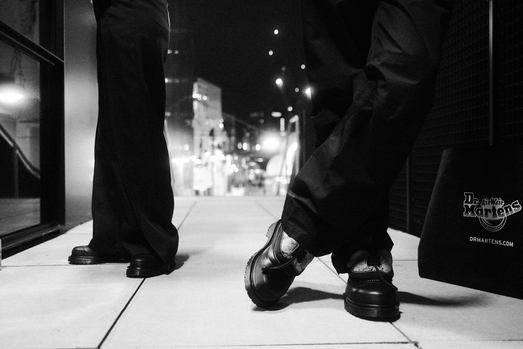 Close-up of two people sitting on a bench at night, with only their legs and feet visible. One person is wearing dark pants and boots, while the other is wearing lighter pants and sneakers. A shopping bag with a logo is beside one person, and city li