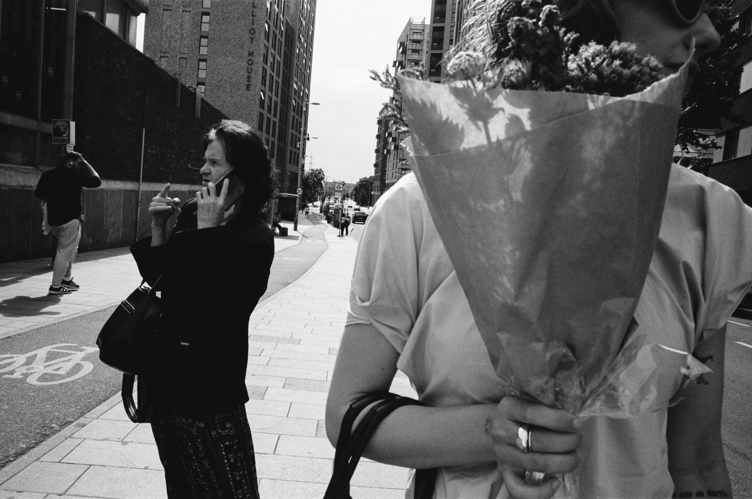 Black-and-white photo showing a woman on her phone, a person holding a bouquet of flowers, and others walking on a city sidewalk with tall buildings in the background.