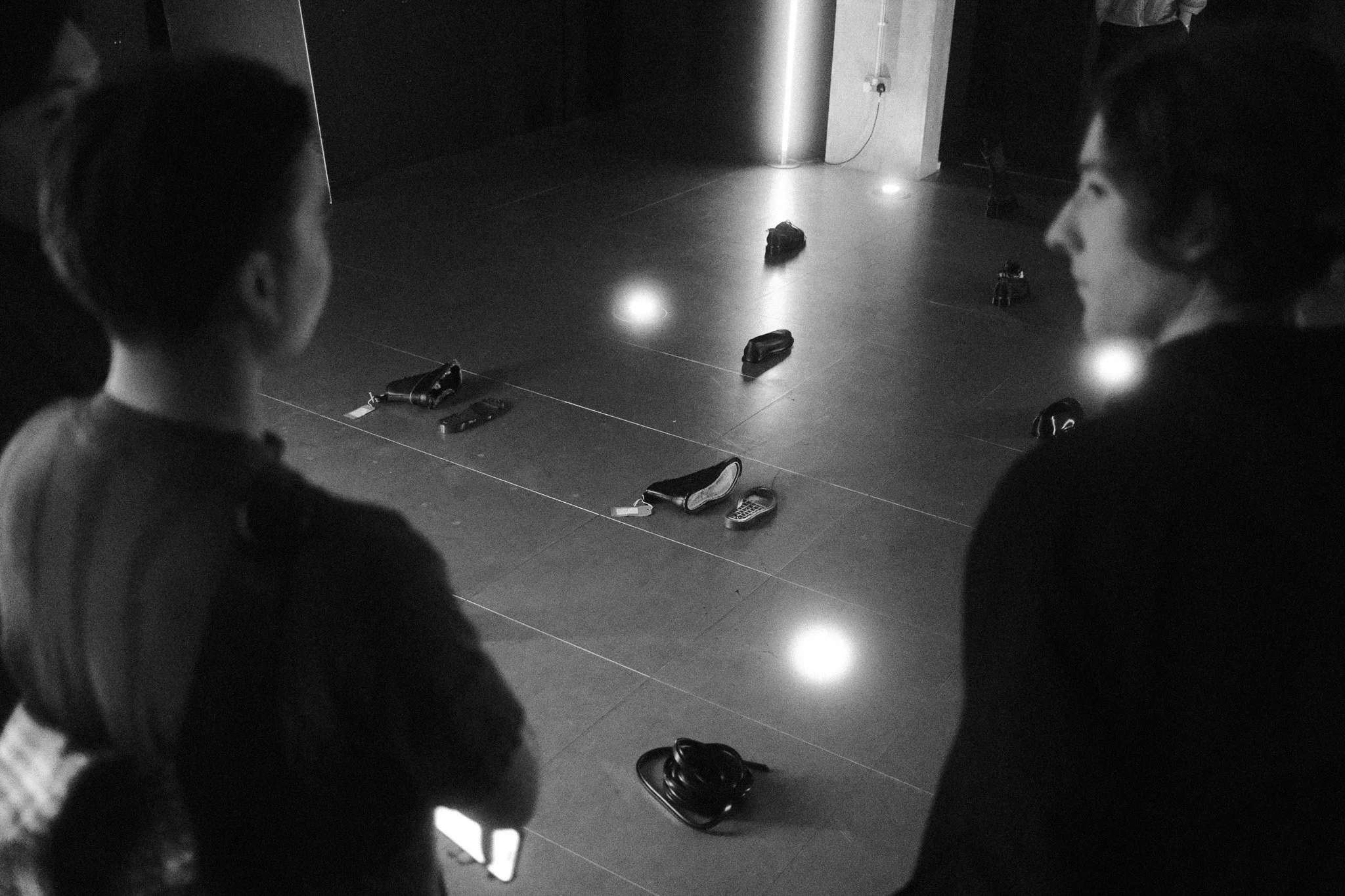 Two people with dark hair are facing each other in front of a floor covered with scattered shoes. The scene is in a dimly lit indoor space, possibly a building lobby or similar area.
