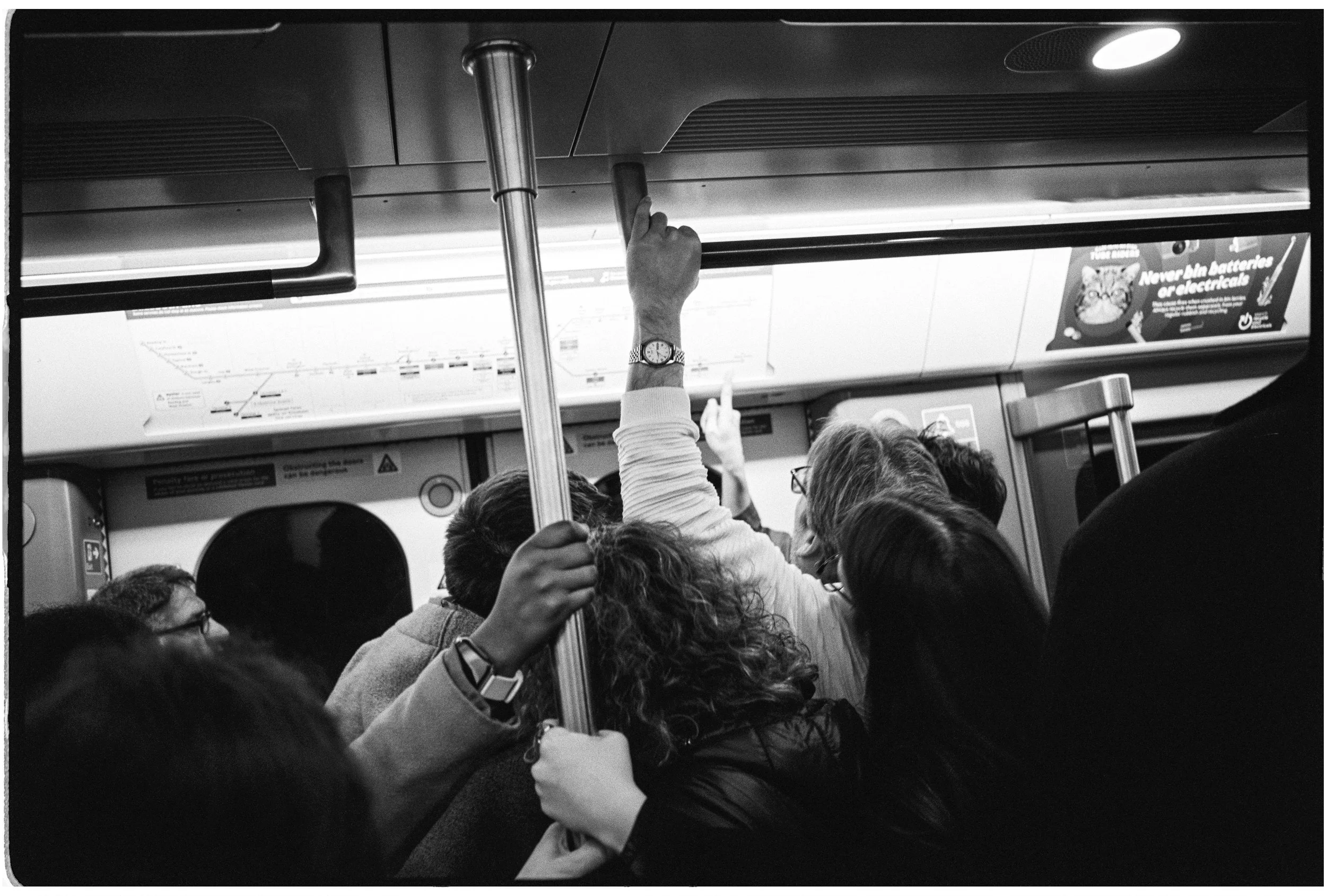 Passengers on a subway train holding onto handrails and poles, with a visible map and advertisement in the background.