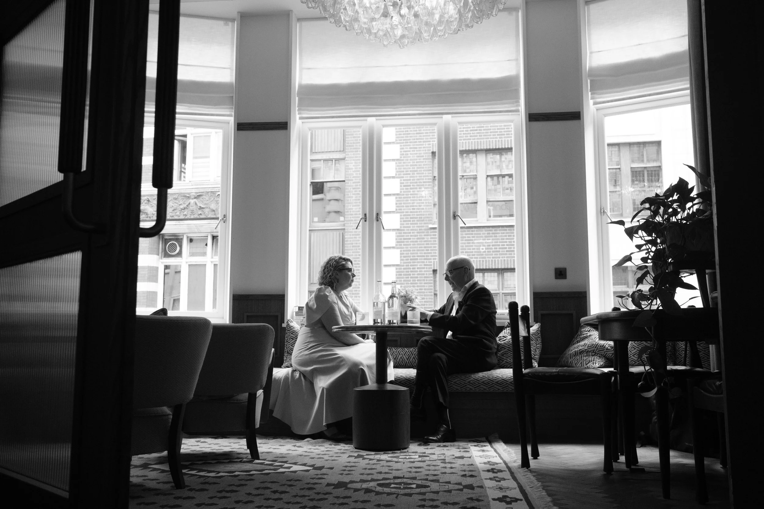 Black and white photo of a couple sitting at a table in a cafe, facing each other and holding hands. Large windows behind them reveal a city view with brick and glass buildings. The ambiance is cozy with a chandelier hanging from the ceiling, and a p