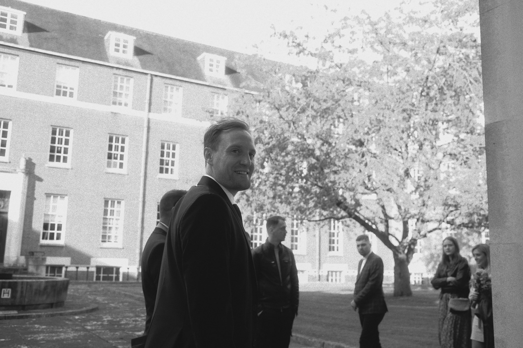 A young man in a suit smiling outdoors with a group of people standing nearby, in front of a large building and trees.