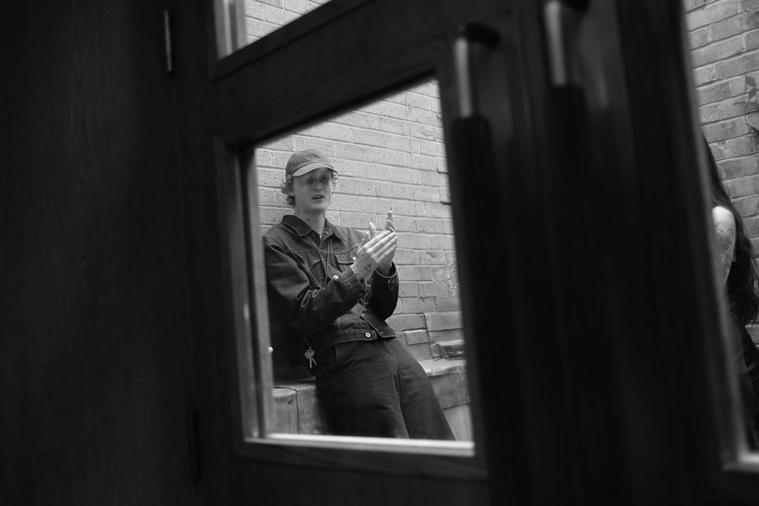 A young person with curly hair and a cap, sitting against a brick wall, captured through a partially opened door or window frame, appearing to be in a moment of attention or conversation.
