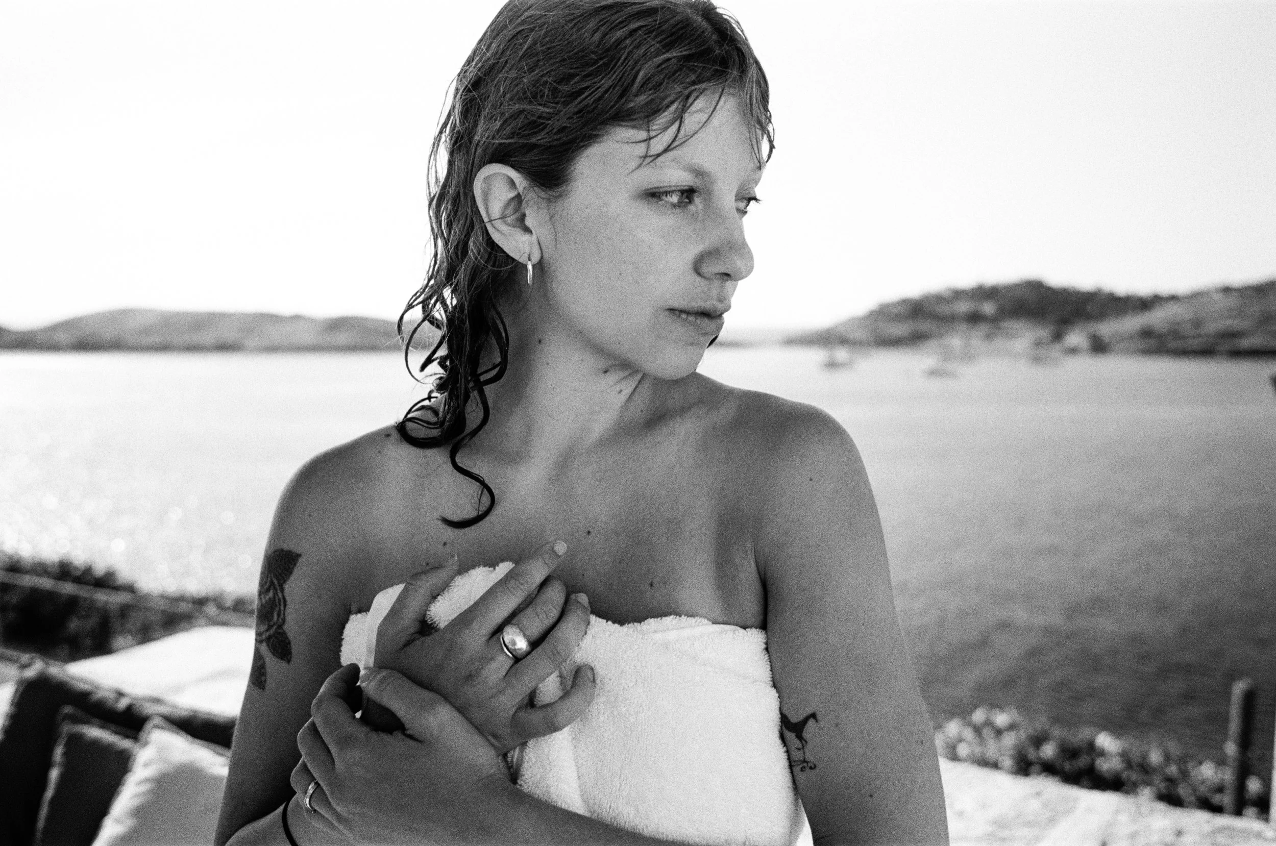 Black and white photograph of a woman with wet hair, wearing earrings and a ring, wrapped in a towel, standing outdoors near the water with hills in the background.