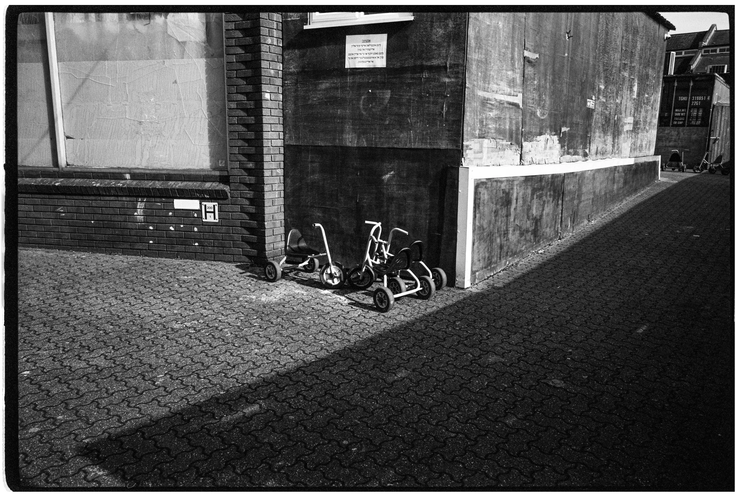 A black and white photo of a city sidewalk with three small tricycles parked against a brick and concrete wall.