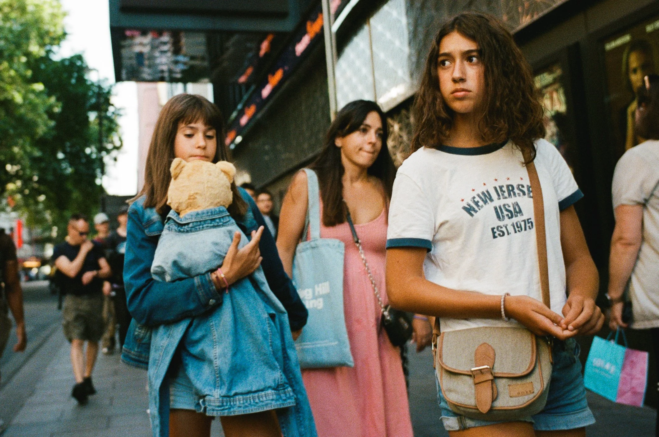 Three young women standing in line outdoors, with one holding a teddy bear, on a city street with trees and buildings in the background.