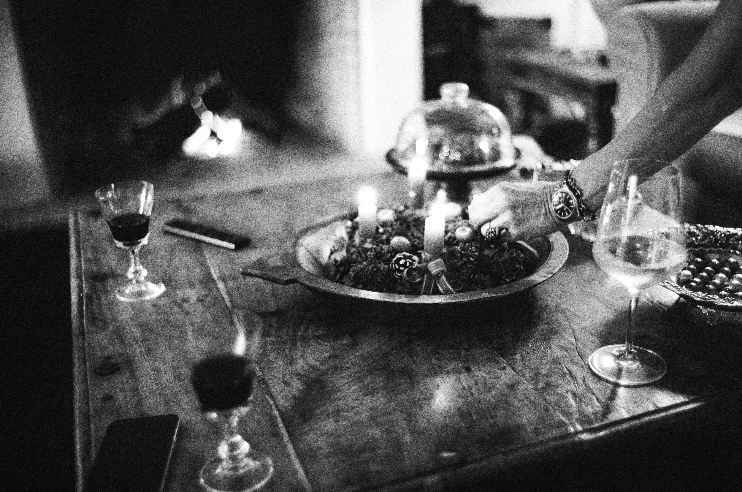 Black and white photo of a tabletop with candles, glasses, and a dish with a decorative centerpiece. A hand is reaching for something on the table, and there are other objects like a small glass and a plate.