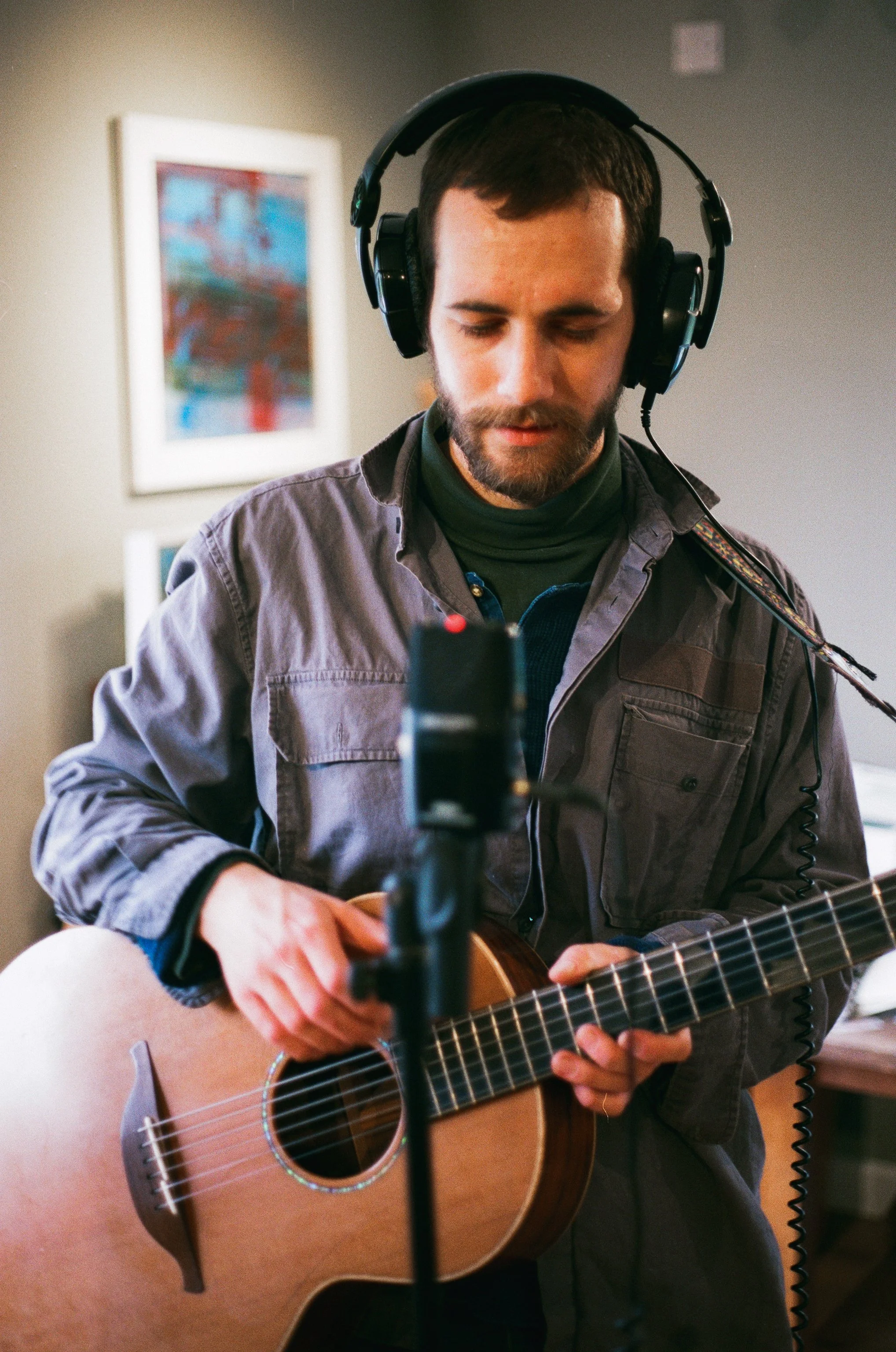A man with short dark hair, beard, and wearing a gray jacket and a green turtleneck is playing an acoustic guitar. He is wearing large black headphones and standing in a room with abstract artwork on the wall. There is a microphone in front of him.