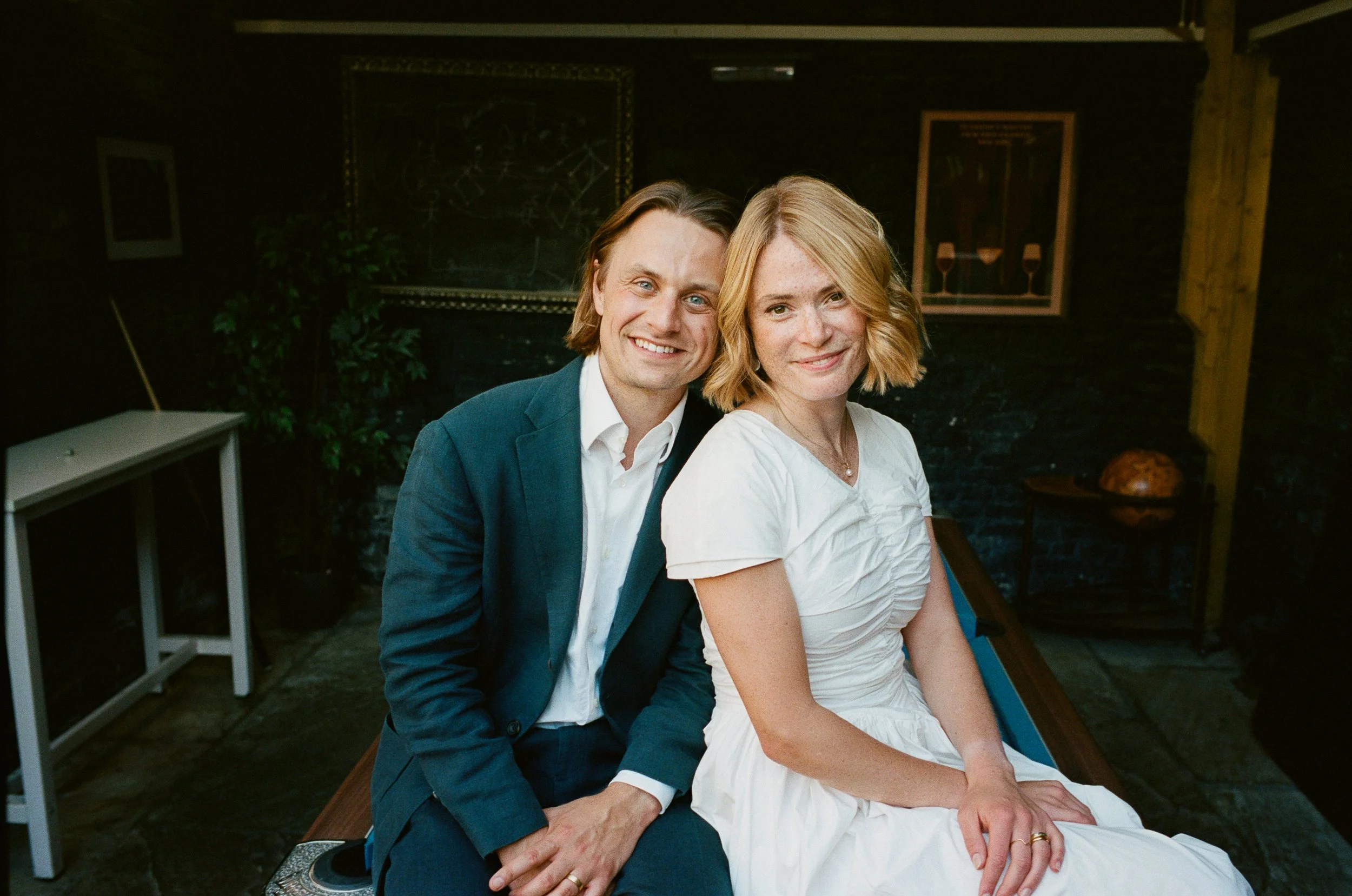 A man and a woman sitting close together, smiling at the camera, in a cozy indoor setting with dark walls, plants, and framed artwork.