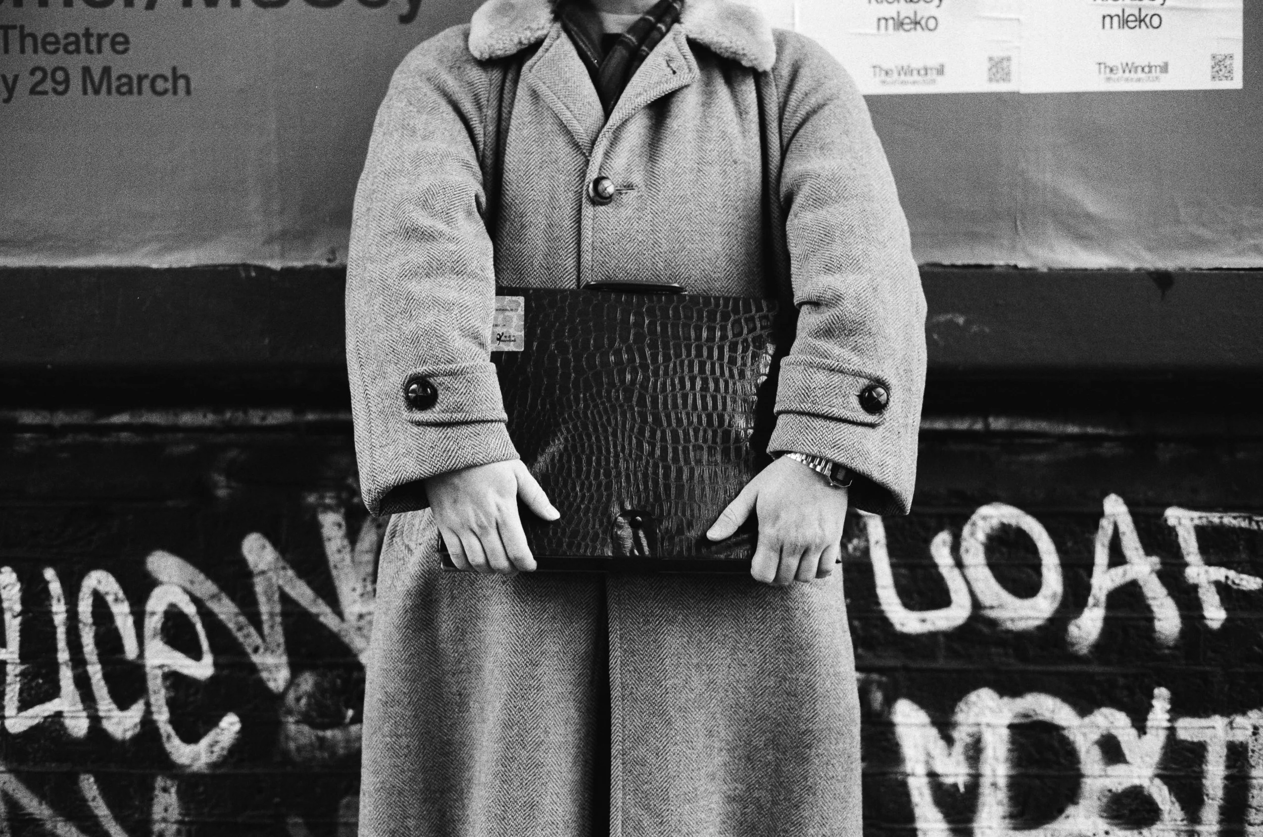Black and white photo of a person wearing a coat with their arms crossed, standing in front of graffiti on a wall.