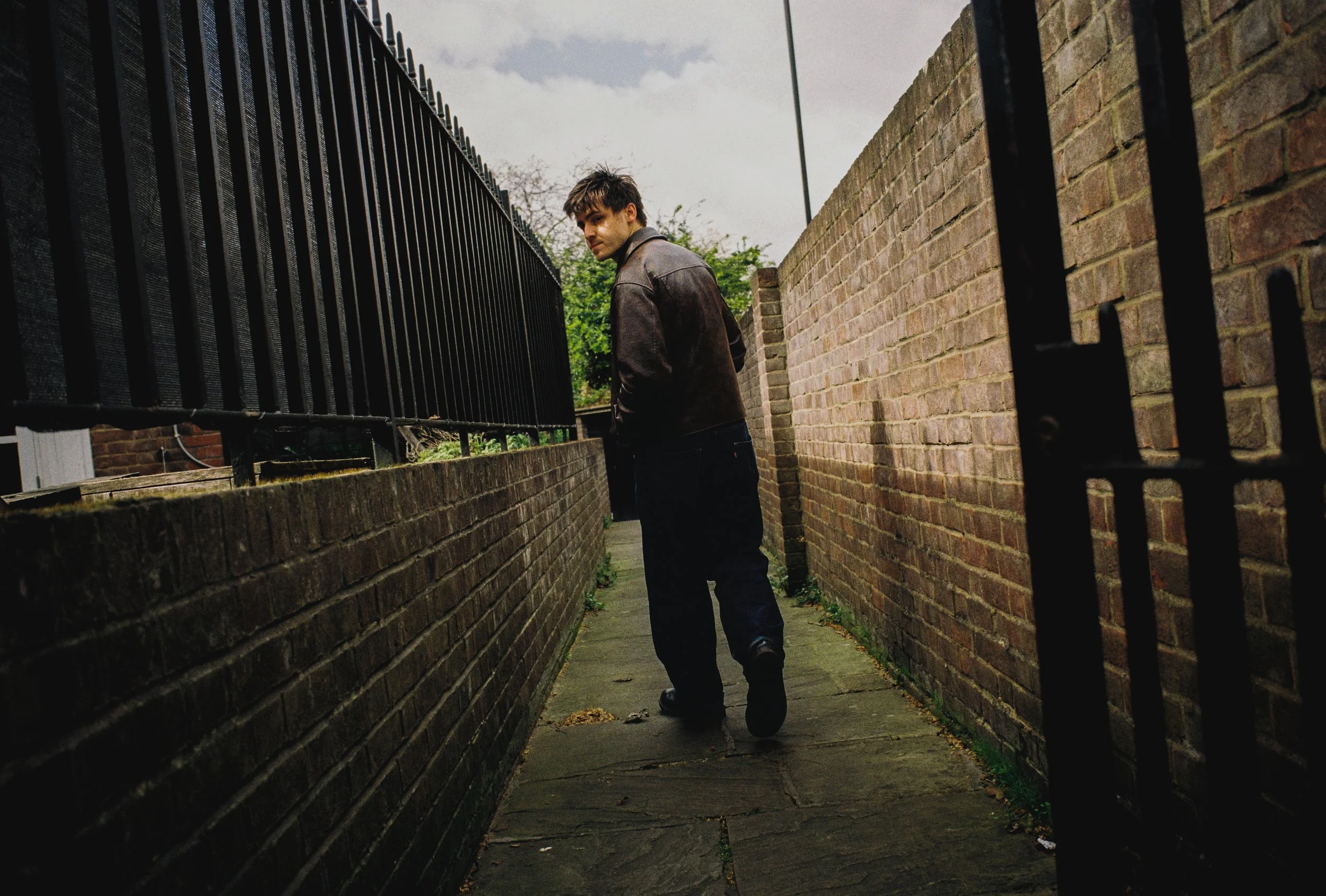 A young man with dark hair wearing a brown leather jacket and dark pants walking down a narrow alley between brick walls, with a black iron fence on his left and a brick wall on his right, under a cloudy sky.