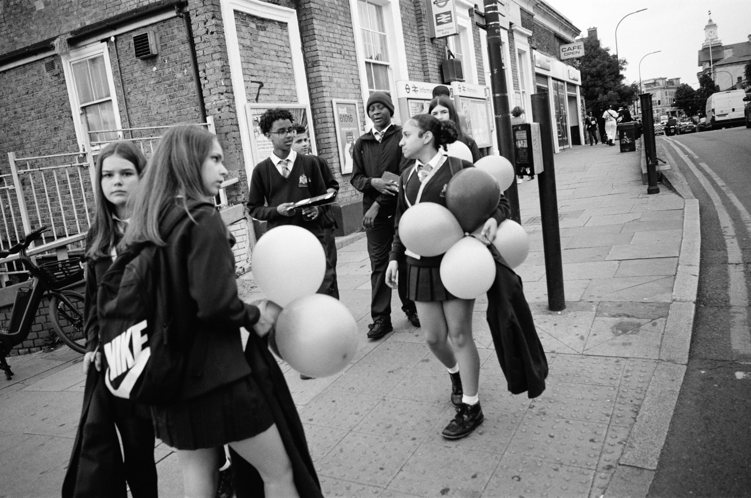 Group of schoolchildren standing on a city sidewalk, some holding balloons, near a bus stop and a brick building.