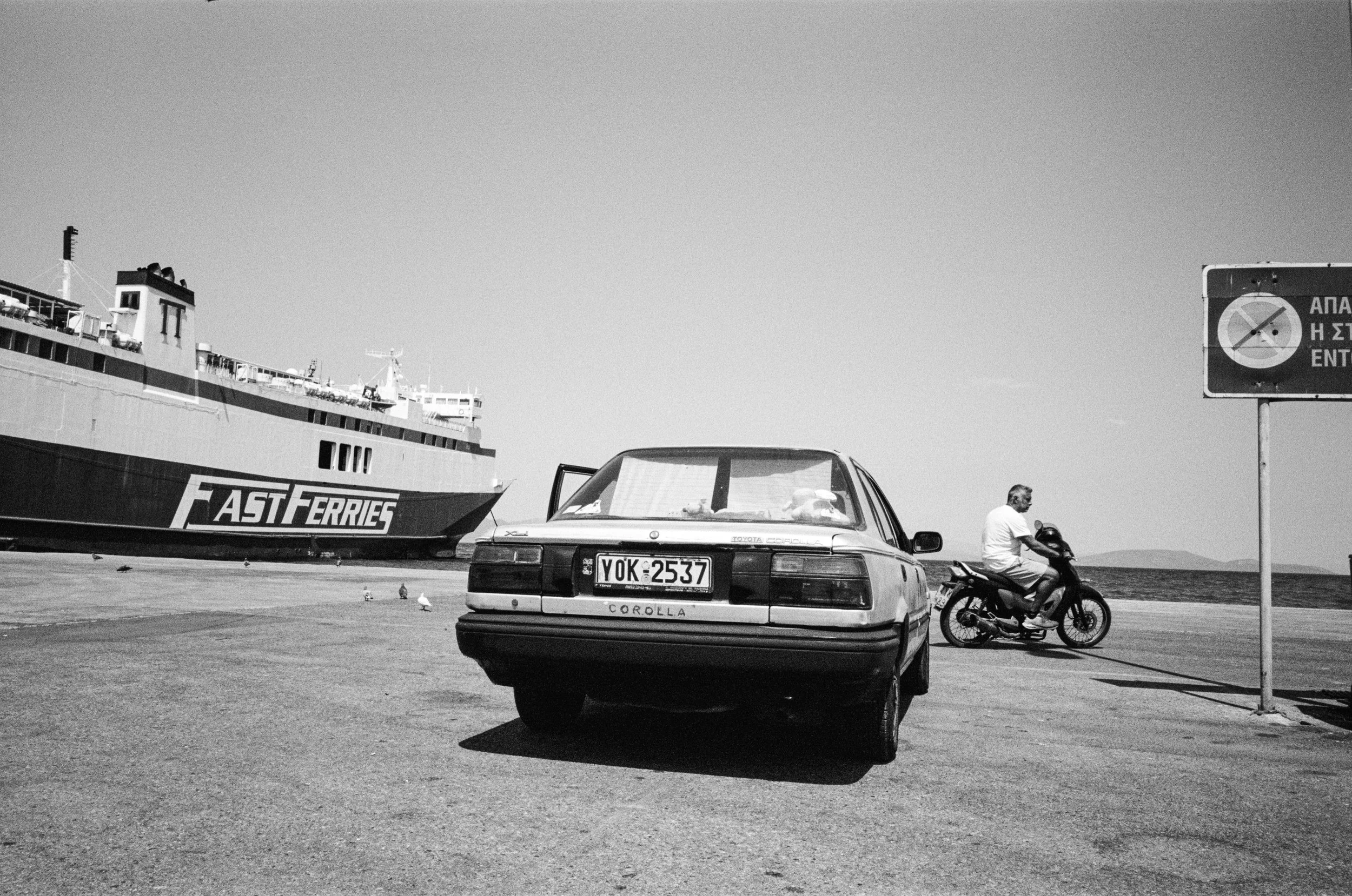 A black and white photo showing a large ferryboat, a parked car with an license plate reading YOK 2537, and a person riding a motorcycle on a road by the seashore. There is a sign with Greek writing and a no-entry symbol. The sky is clear and the sce